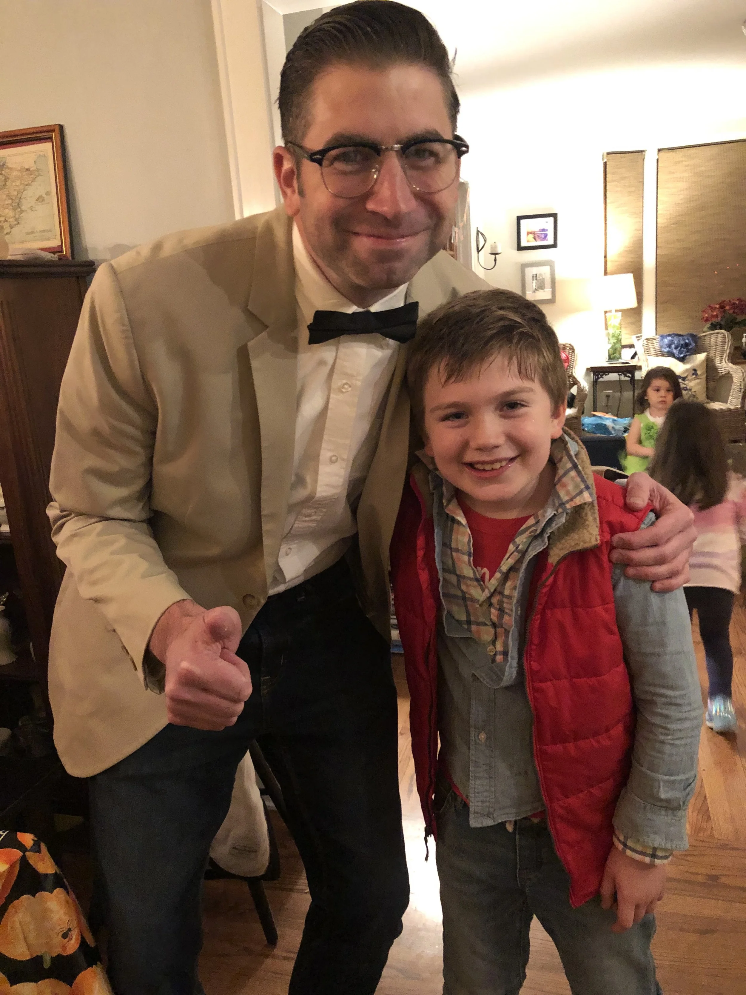 A man in a tan blazer, white shirt, and black bow tie smiling and giving a thumbs-up, standing next to a boy in a red vest and plaid shirt, both smiling and posing for the camera inside a warmly lit room with children in the background.