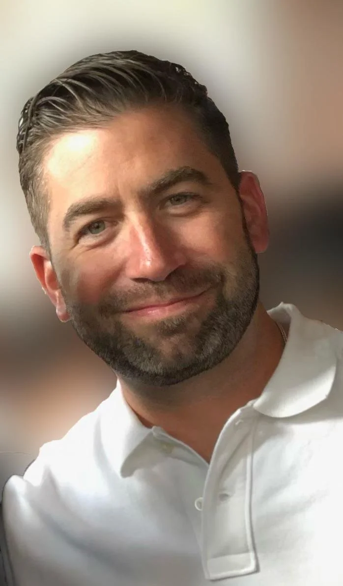Close-up of a smiling man with dark hair, light skin, and a beard, wearing a white collared shirt.