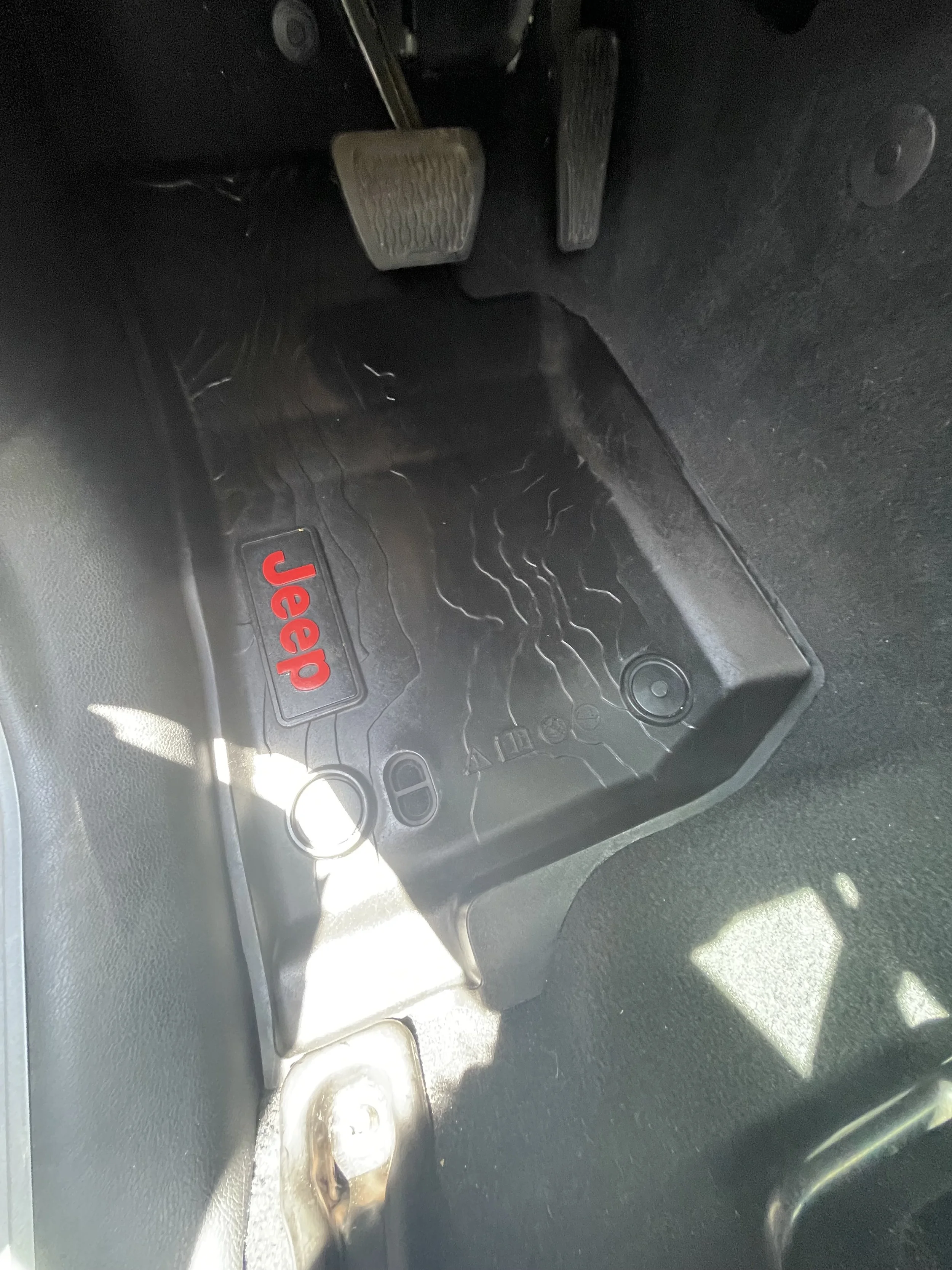 The image shows the foot pedals of a Jeep vehicle, including the accelerator, brake, and clutch pedals, with a Jeep-branded floor mat underneath.