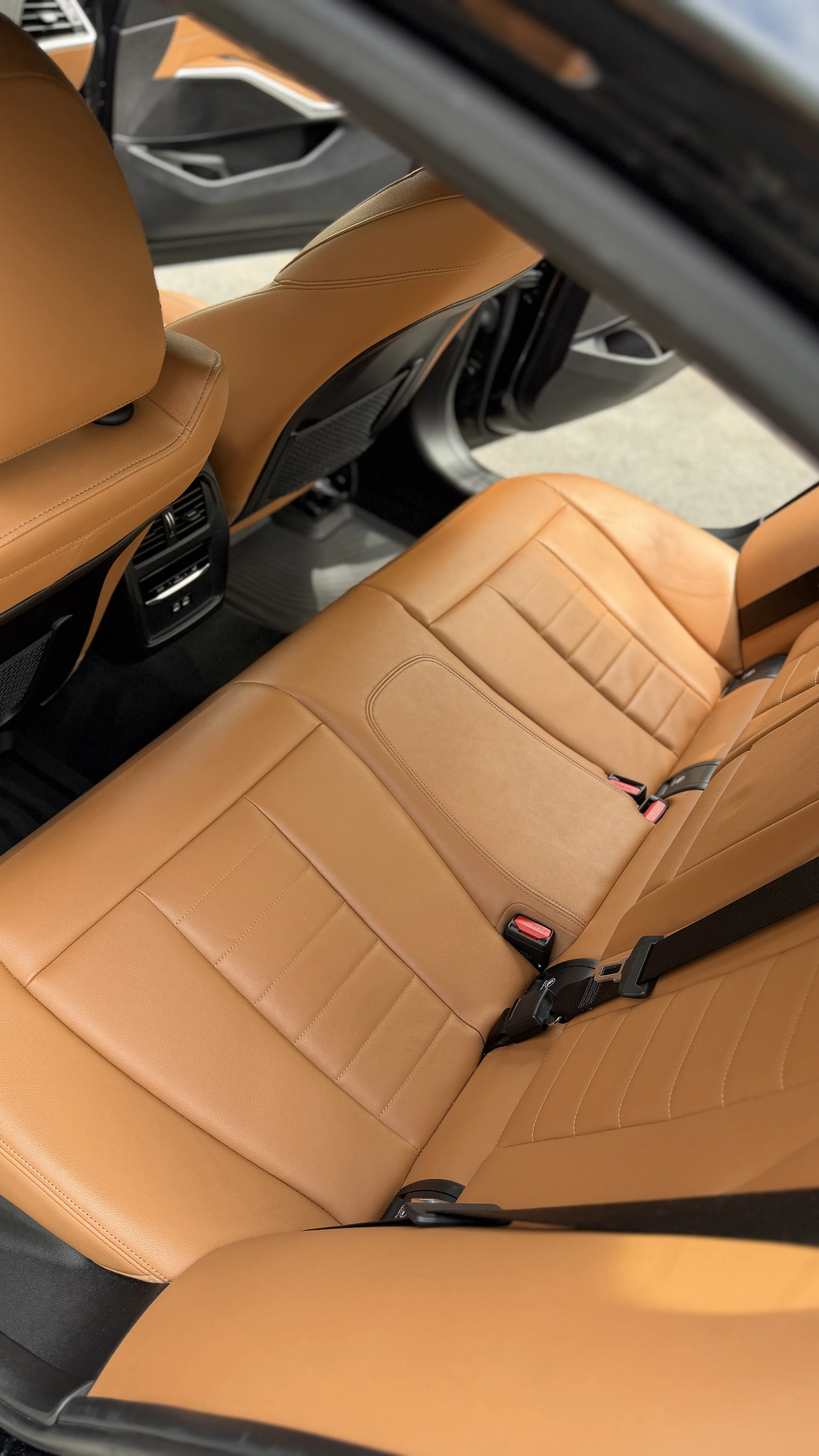 Back seat of a car with tan leather seats and black seat belts.