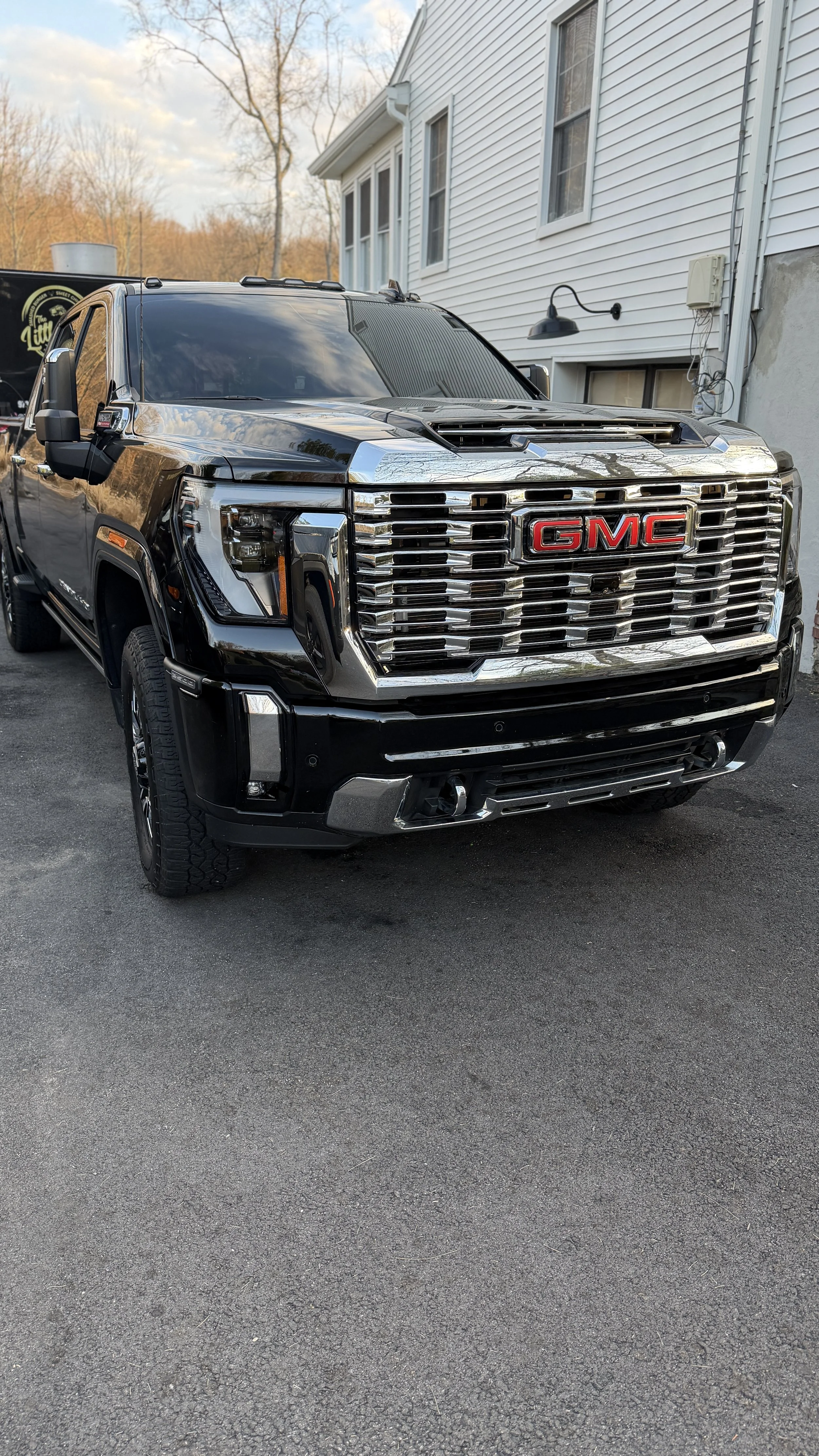 A shiny black GMC pickup truck parked on a driveway beside a white house with vinyl siding, with trees and a partly cloudy sky in the background.