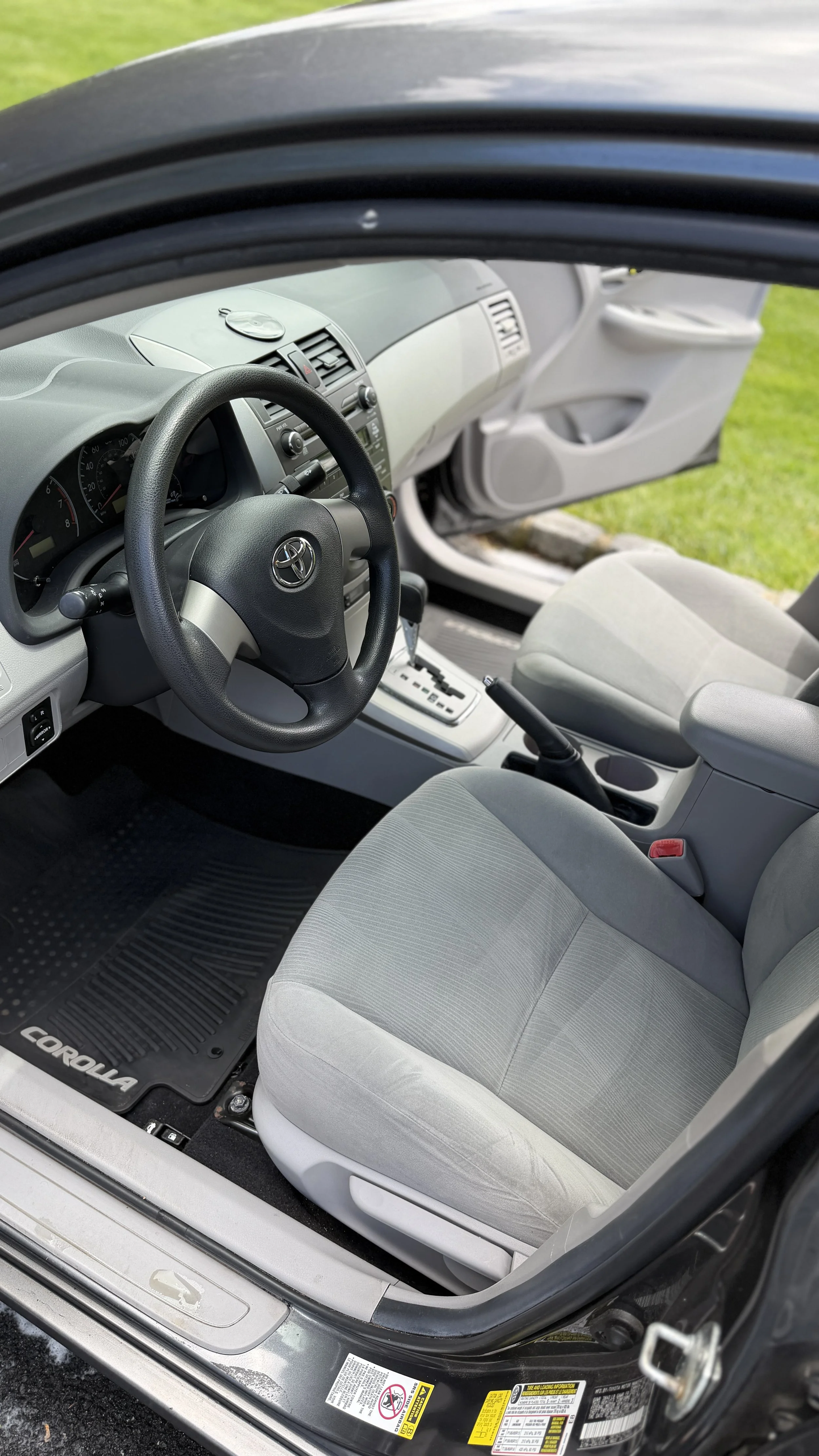 The interior of a Toyota Corolla car showing the steering wheel, front seats, dashboard, and open driver's side door with grass outside.