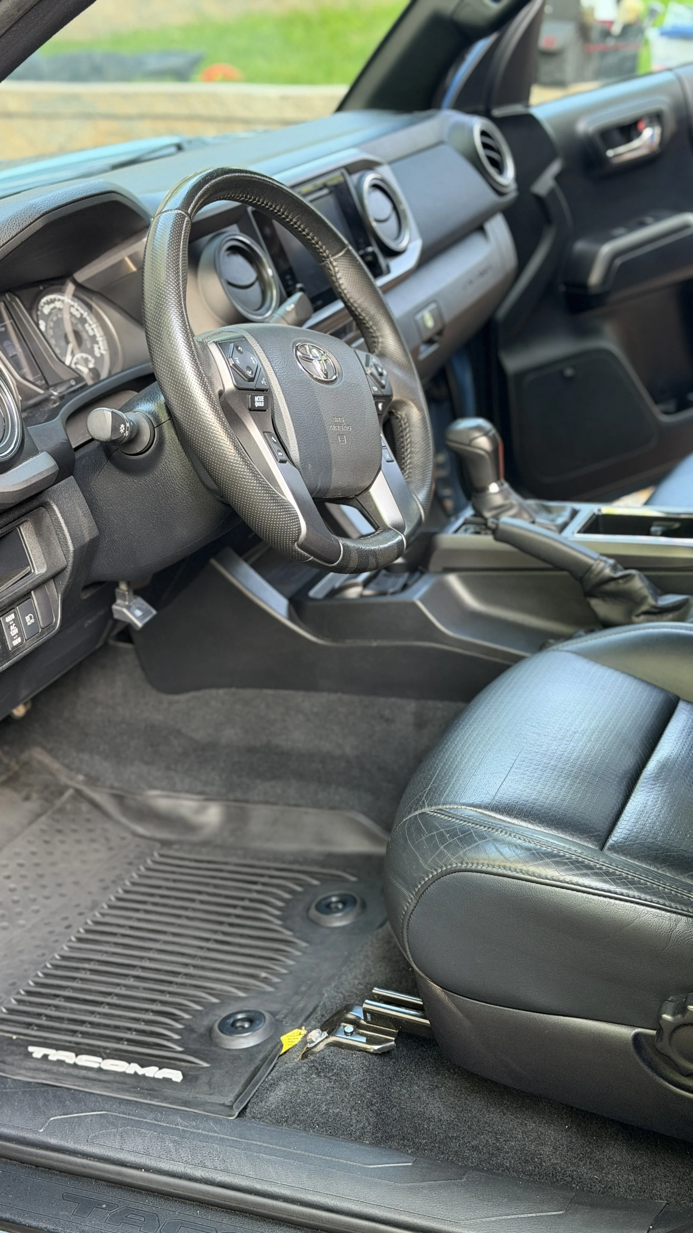 Interior view of a Toyota Tacoma truck's driver side showing a black leather seat, steering wheel with controls, dashboard with gauges, gear shift, and floor mat with 'Tacoma' lettering.
