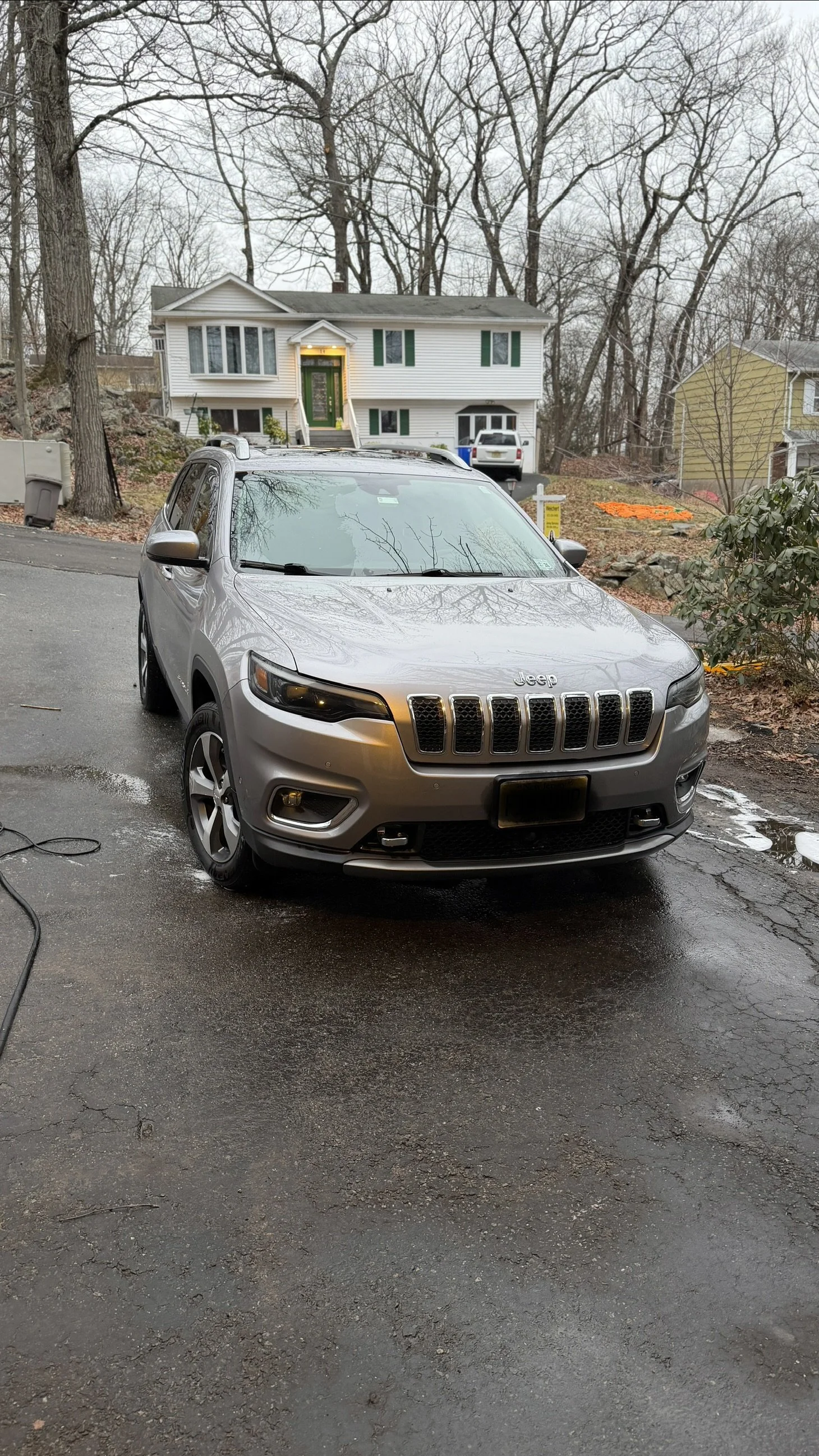 A gray Jeep SUV parked on a wet asphalt driveway in front of a suburban house with a green front door and green shutters on the windows, surrounded by leafless trees and overcast sky.