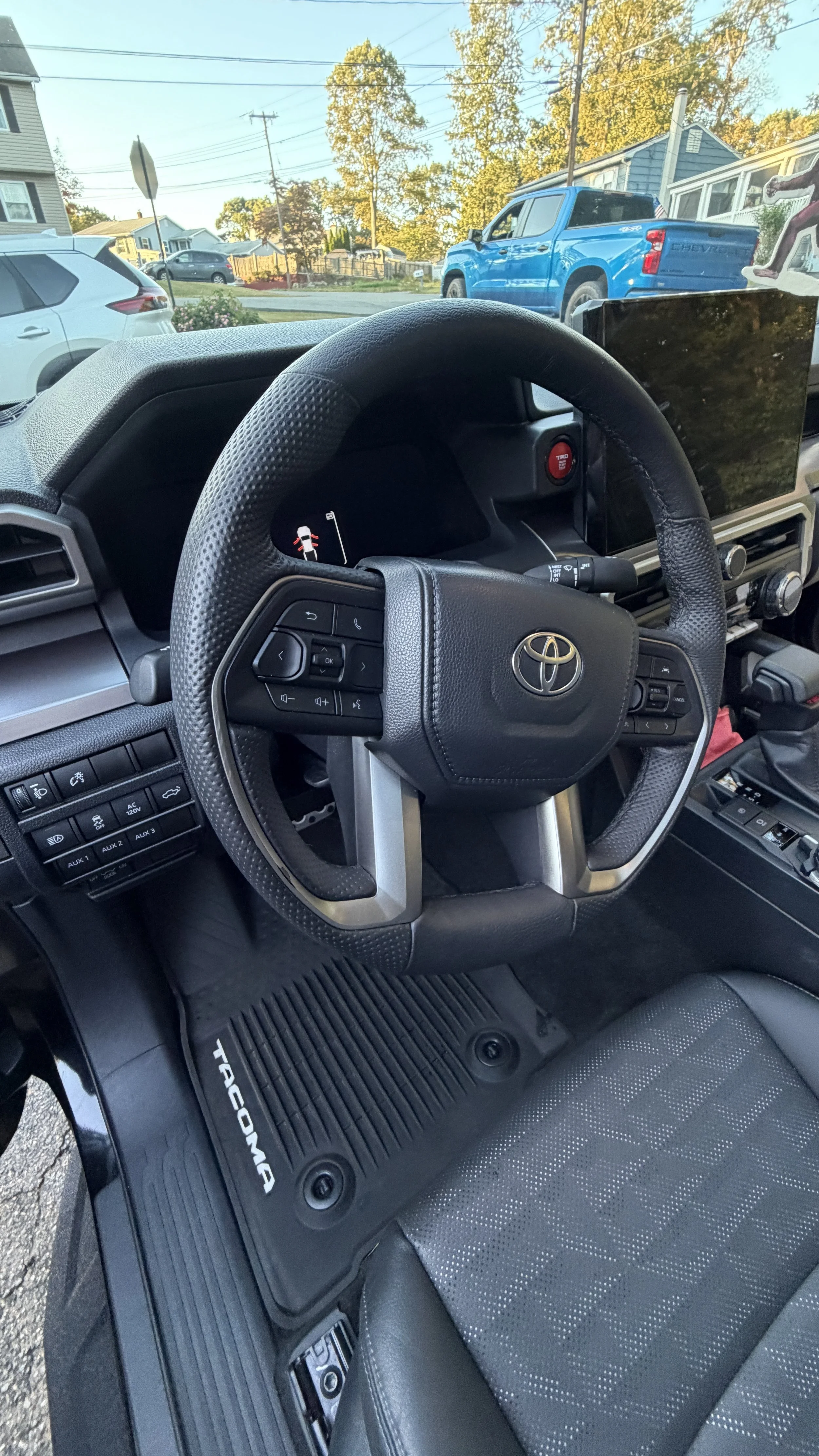 View from inside a Toyota Tacoma truck showing the steering wheel, dashboard, and front passenger seat, with a residential neighborhood and parked cars visible through the windshield.