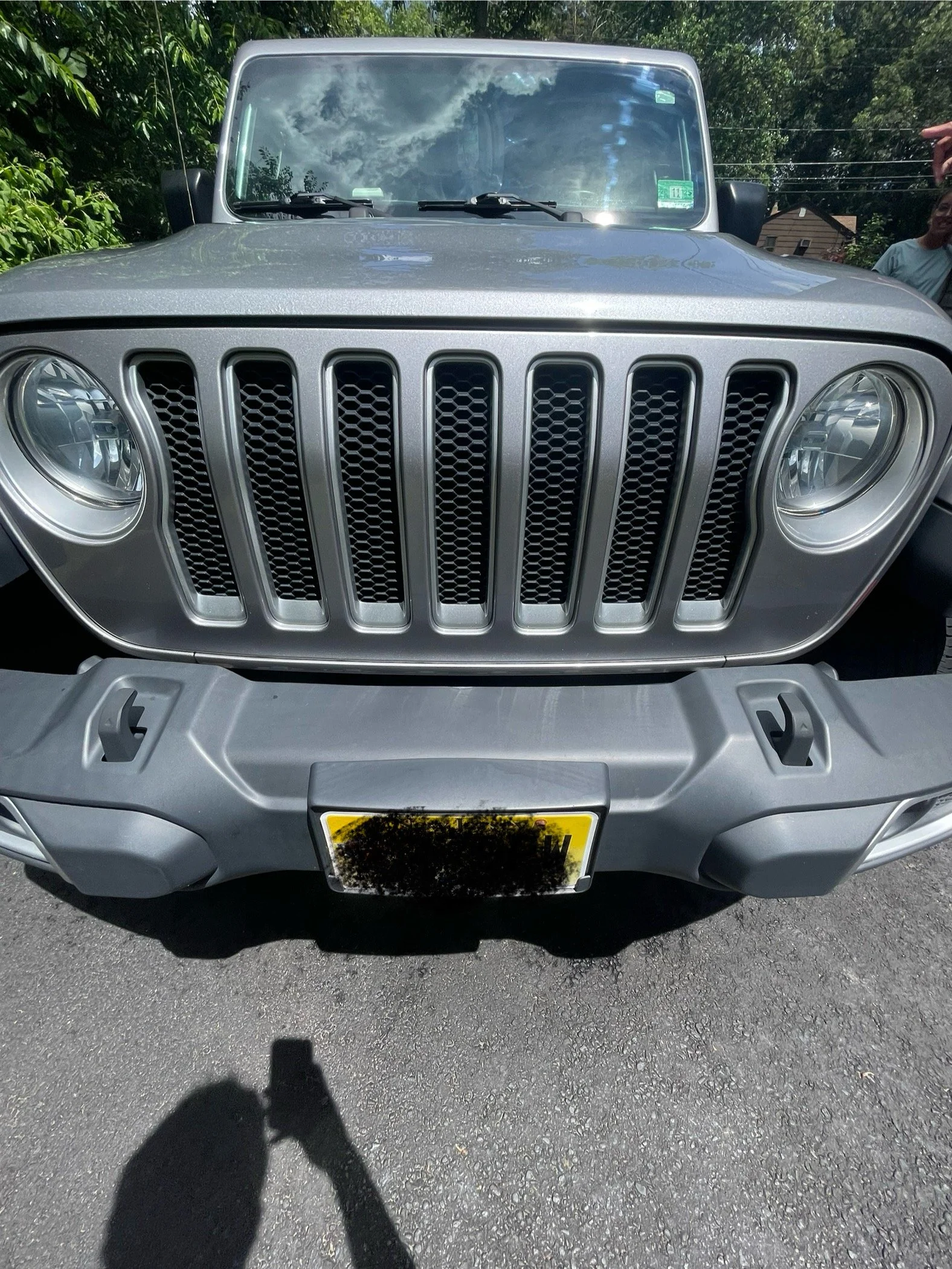 Close-up of the front view of a gray Jeep with a distinctive grille, round headlights, and a portion of the license plate visible, parked on an asphalt surface with trees in the background.