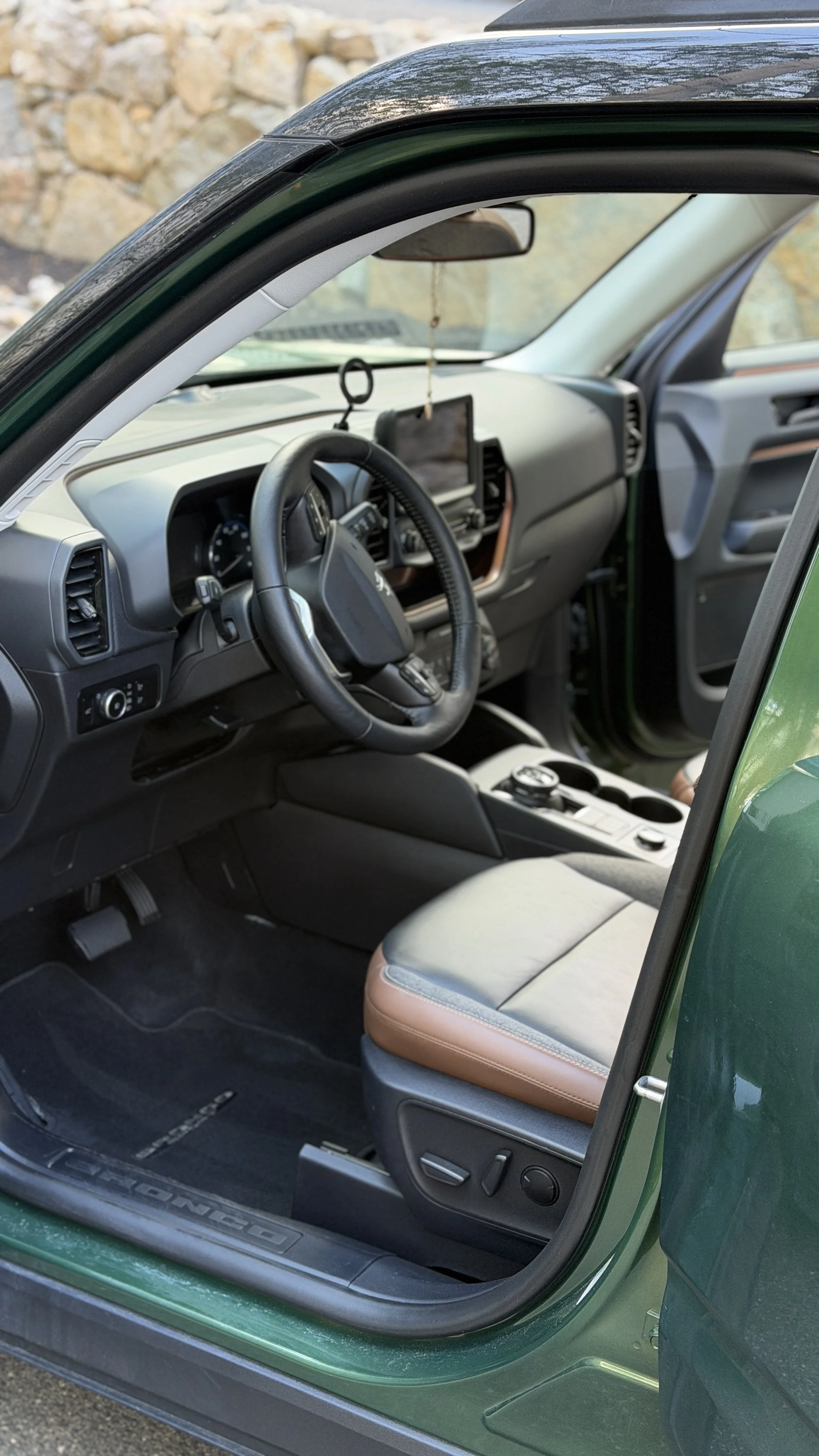 Inside view of a green vehicle with an open door, showing a black steering wheel, dashboard with digital display, center console with controls, and a tan and gray seat.