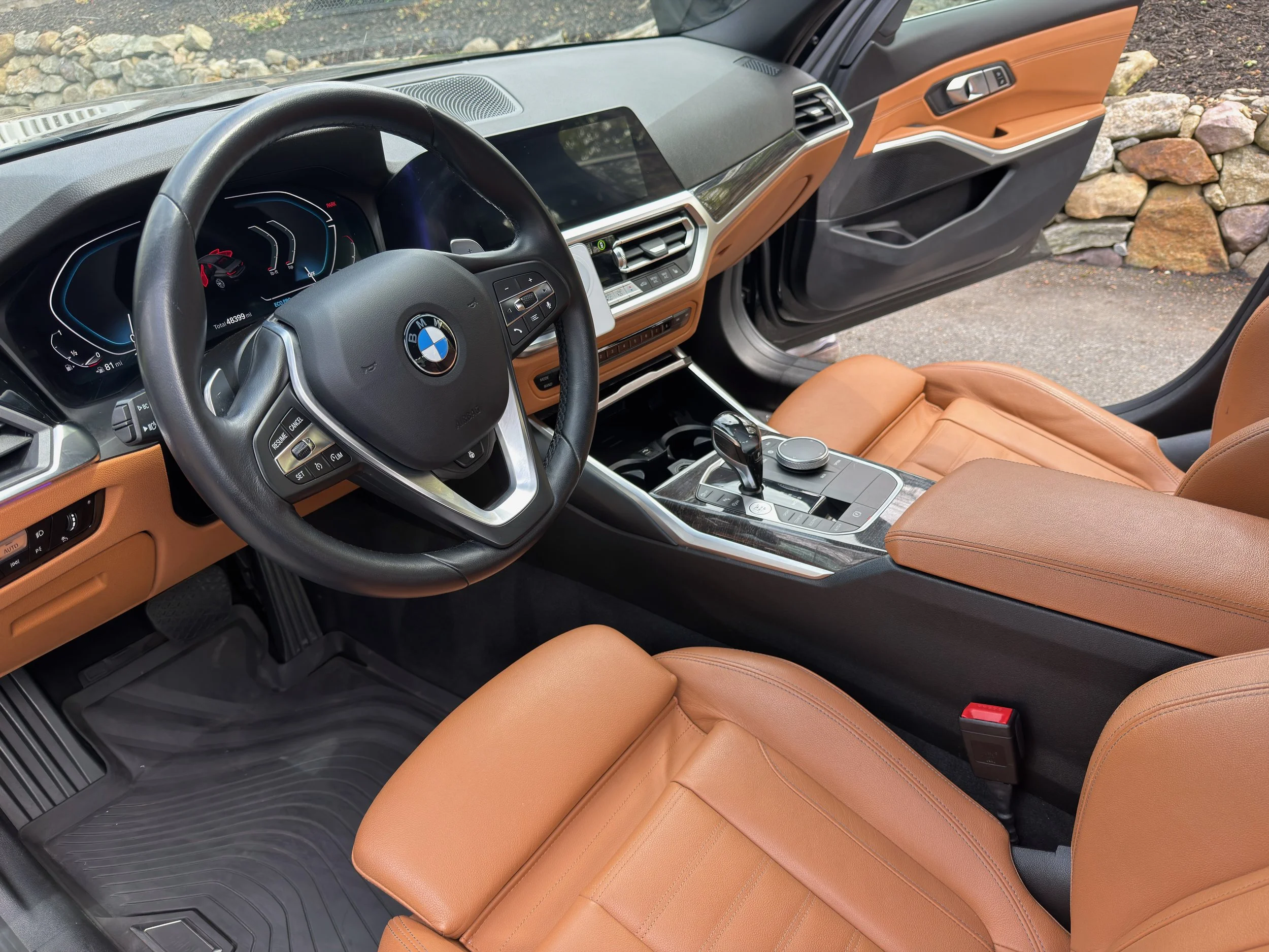 Interior of a BMW vehicle showing the steering wheel, dashboard, center console, and tan leather seats with a stone wall outside the open door.