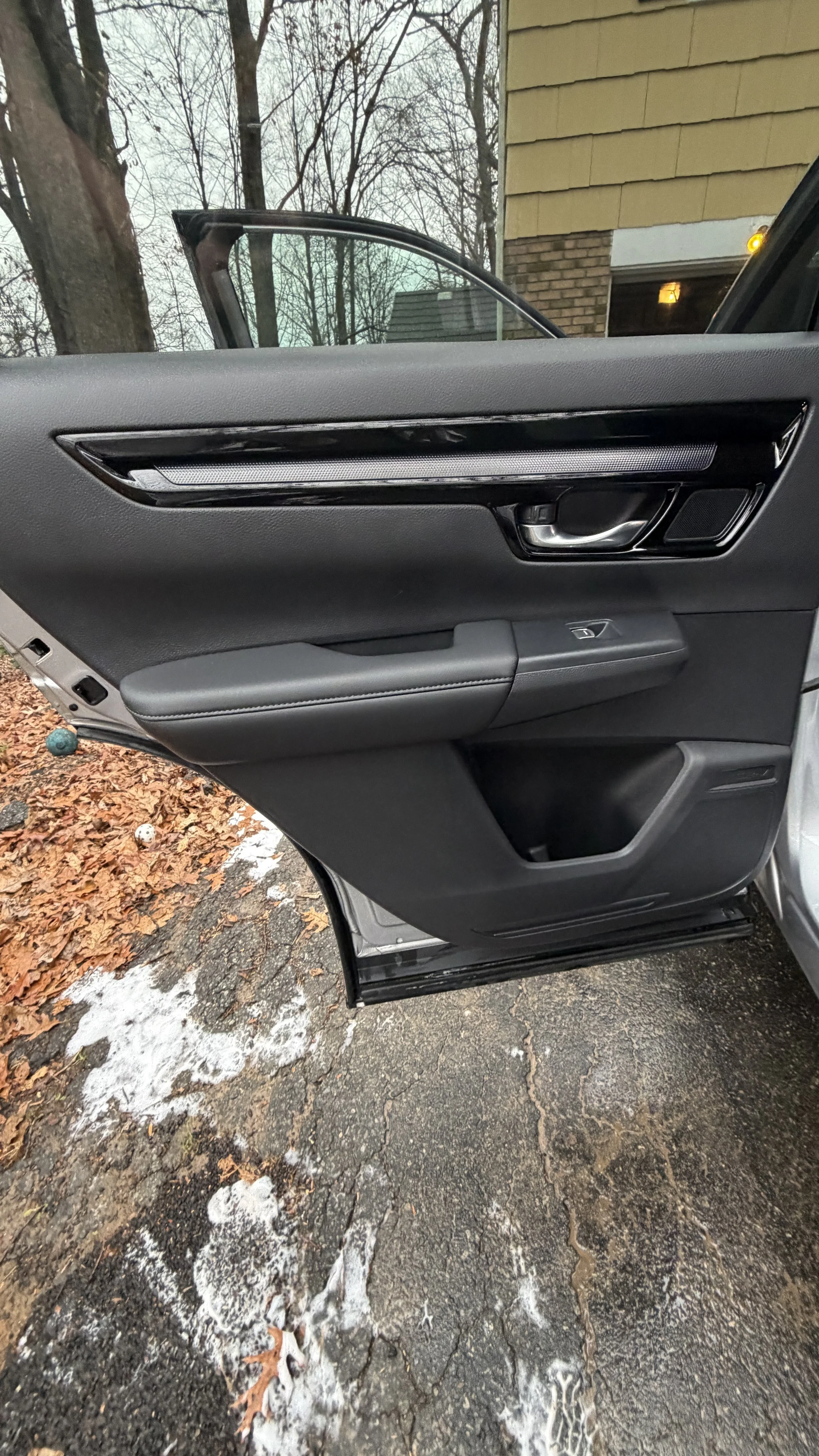 The image shows the inside of a car door with a window partly rolled down. Outside, there are fallen leaves on an asphalt pavement, some patches of snow, and bare trees indicating a late autumn or early winter setting.