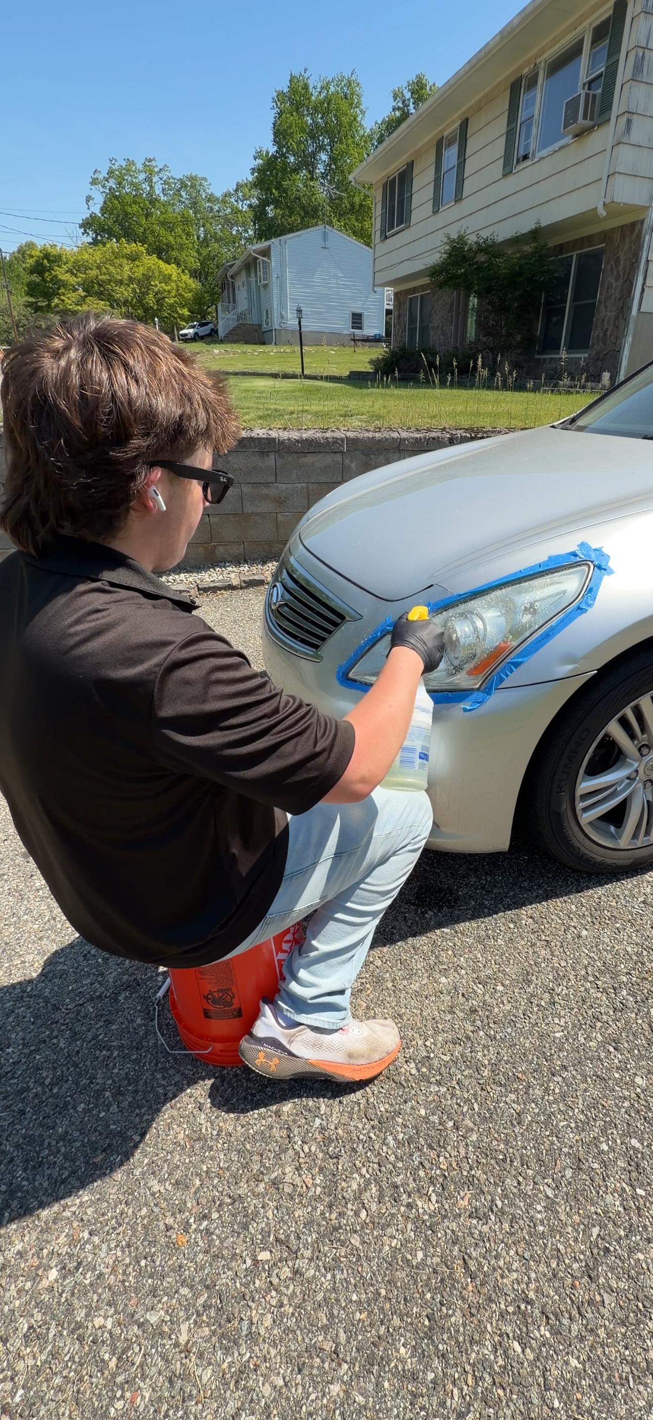 A person wearing glasses and gloves is cleaning the headlight of a white car with a spray bottle. The car is parked outdoors on a paved surface, with a residential house and greenery in the background.