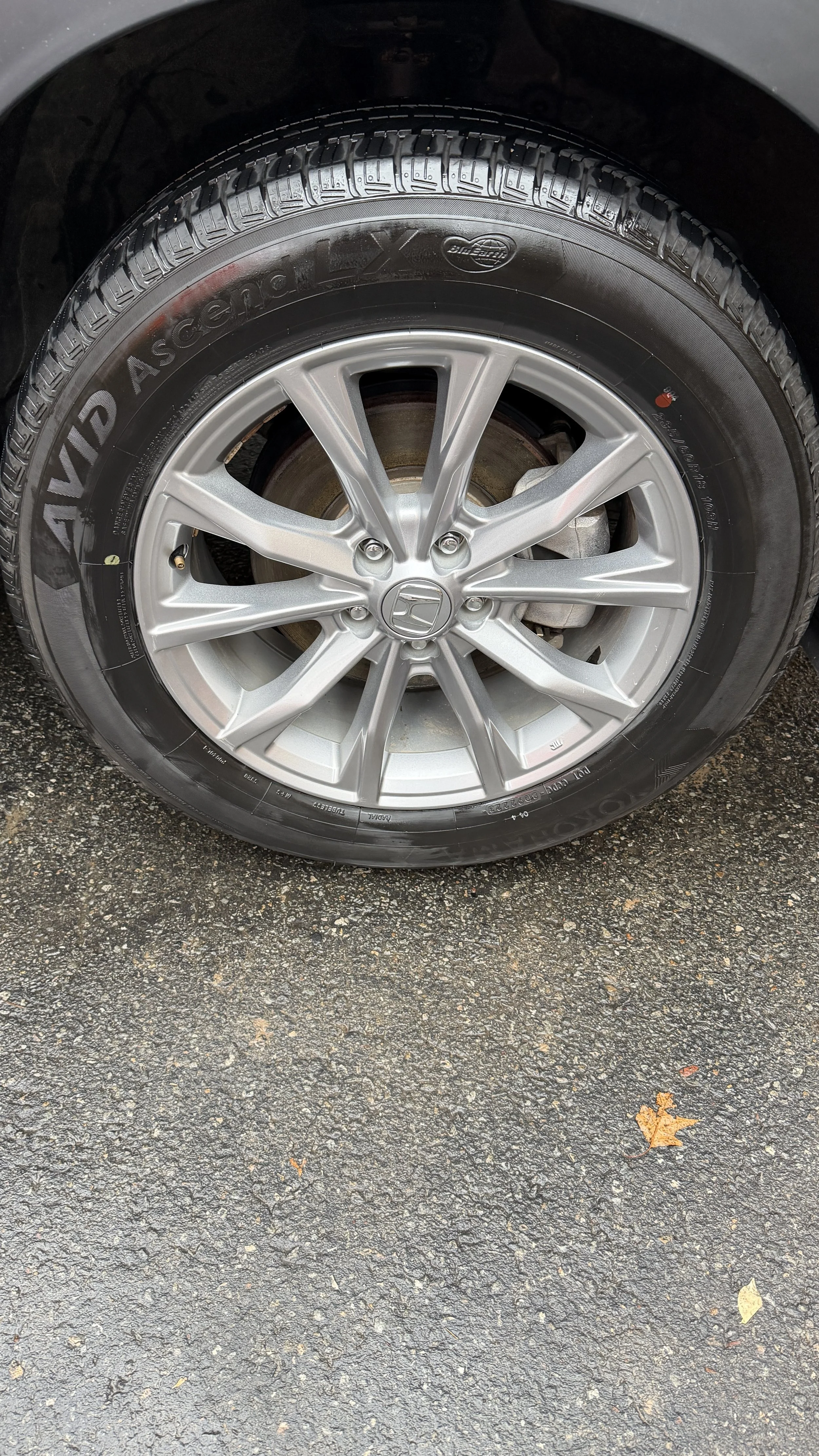 Close-up of a car tire with a silver alloy wheel, the ground is asphalt with some fallen leaves.