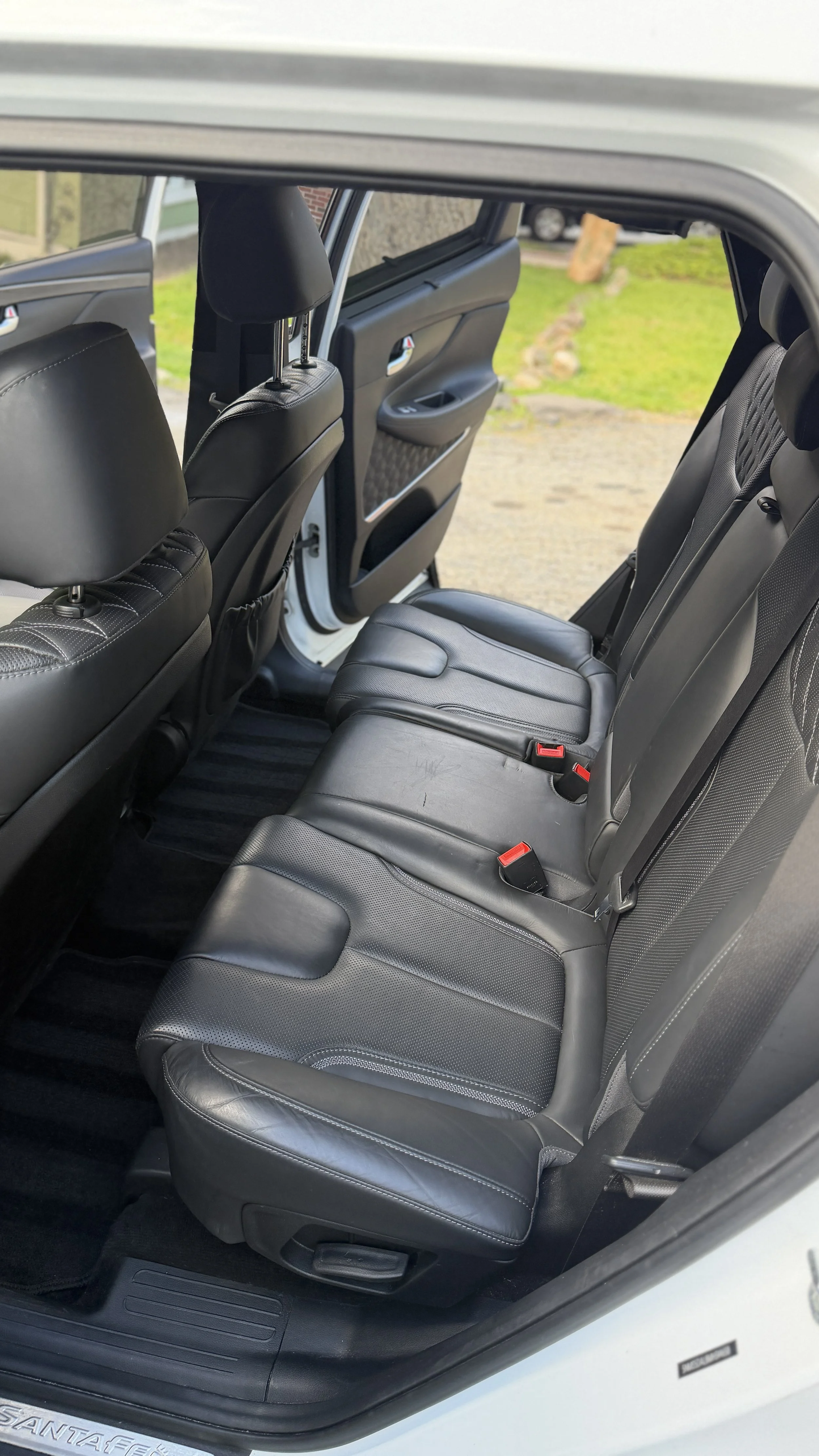 Rear interior of a vehicle showing black leather seats with seat belts, black door panels, and a view of a grassy area outside.