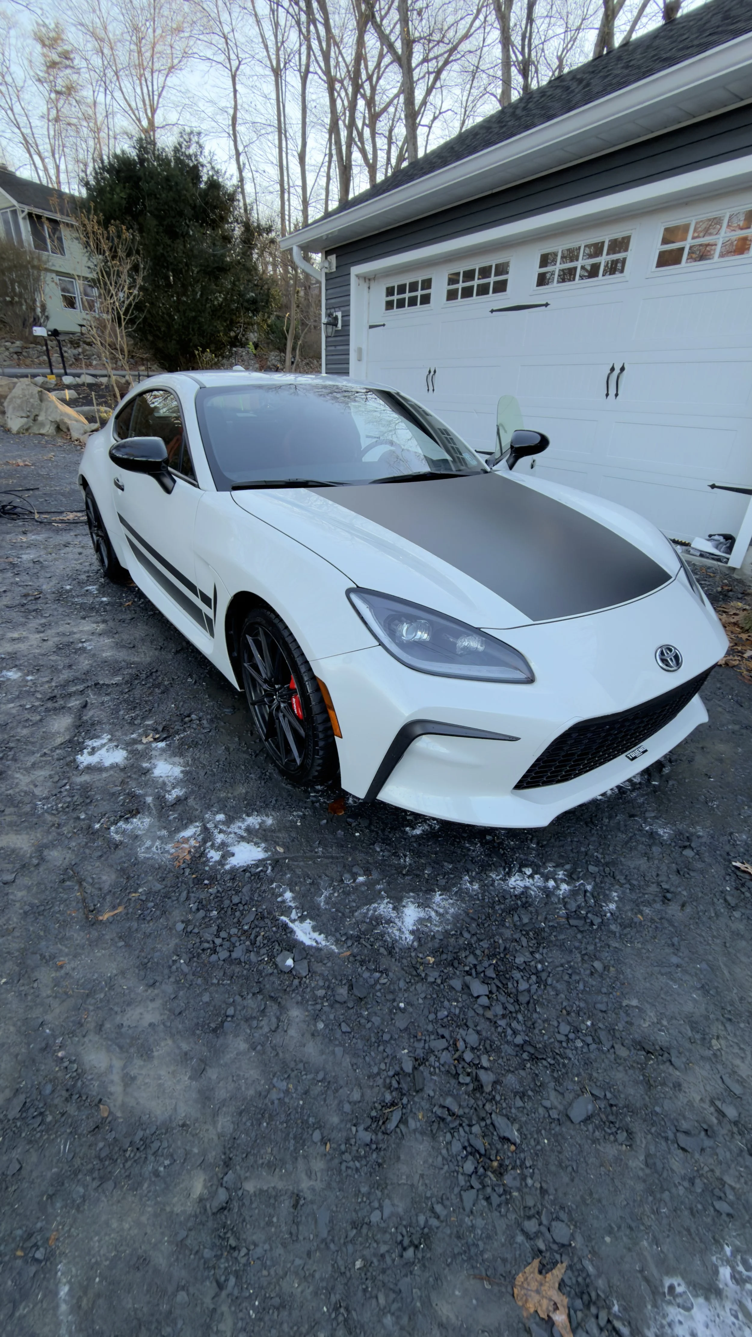 A white sports car parked on a gravel driveway in front of a gray garage door, with a reflection of trees on the car's windshield and hood.
