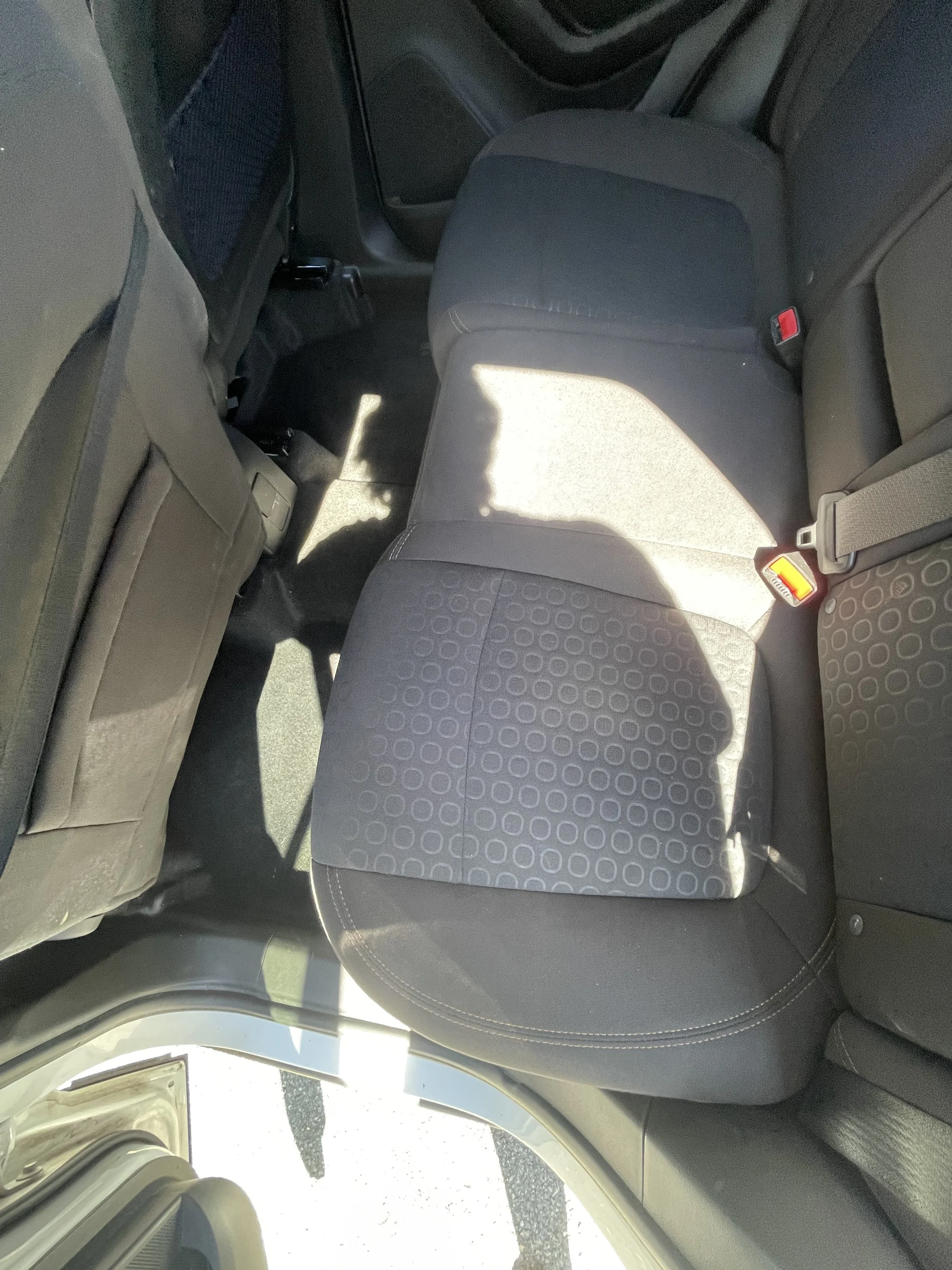 The backseat interior of a car, showing the middle and part of the passenger side seats, both covered in dark fabric with circular pattern, seat belts, and a window partially reflecting light.