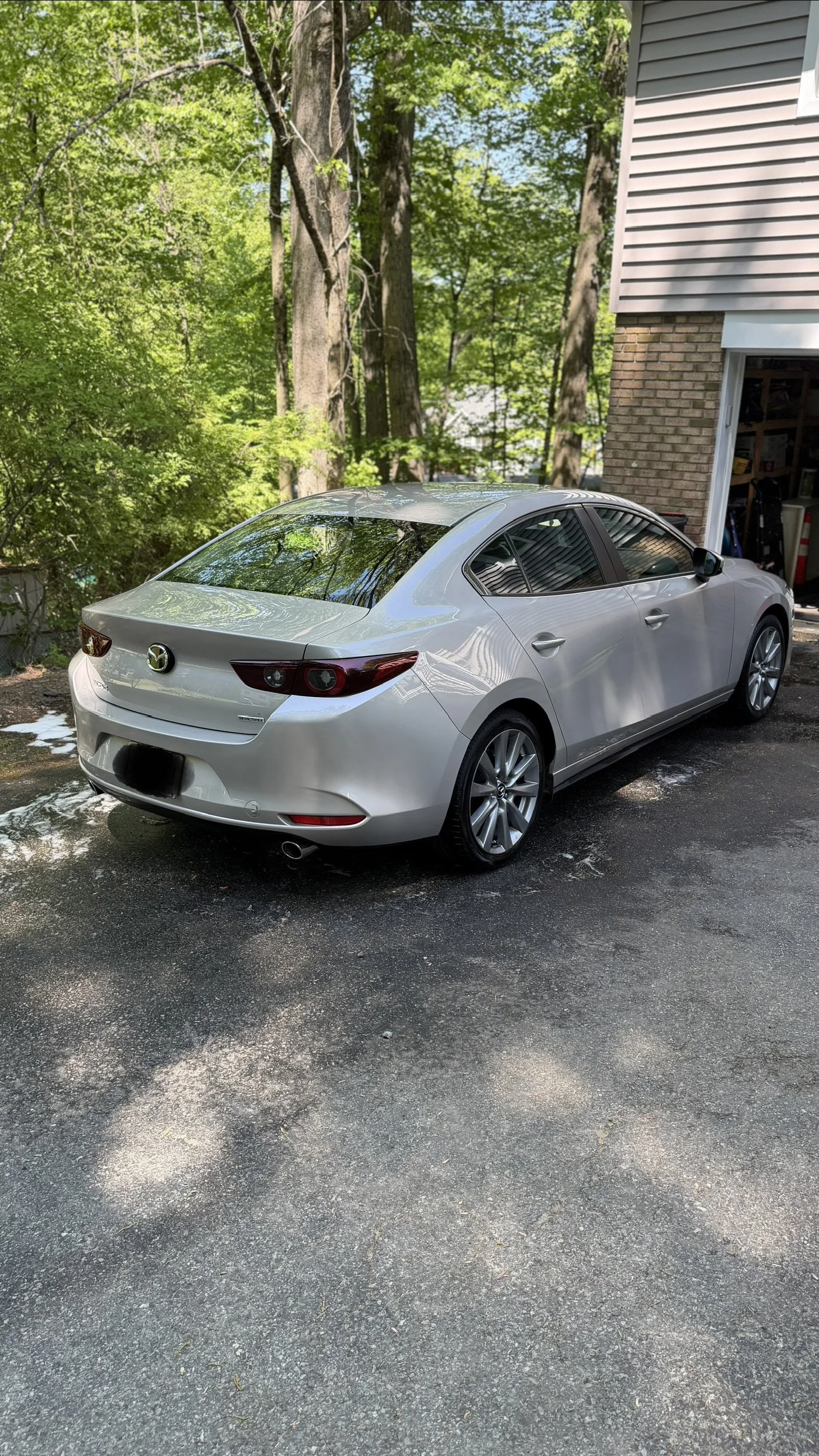 Silver Mazda sedan parked on a residential driveway surrounded by green trees and a house with a brick and siding exterior.
