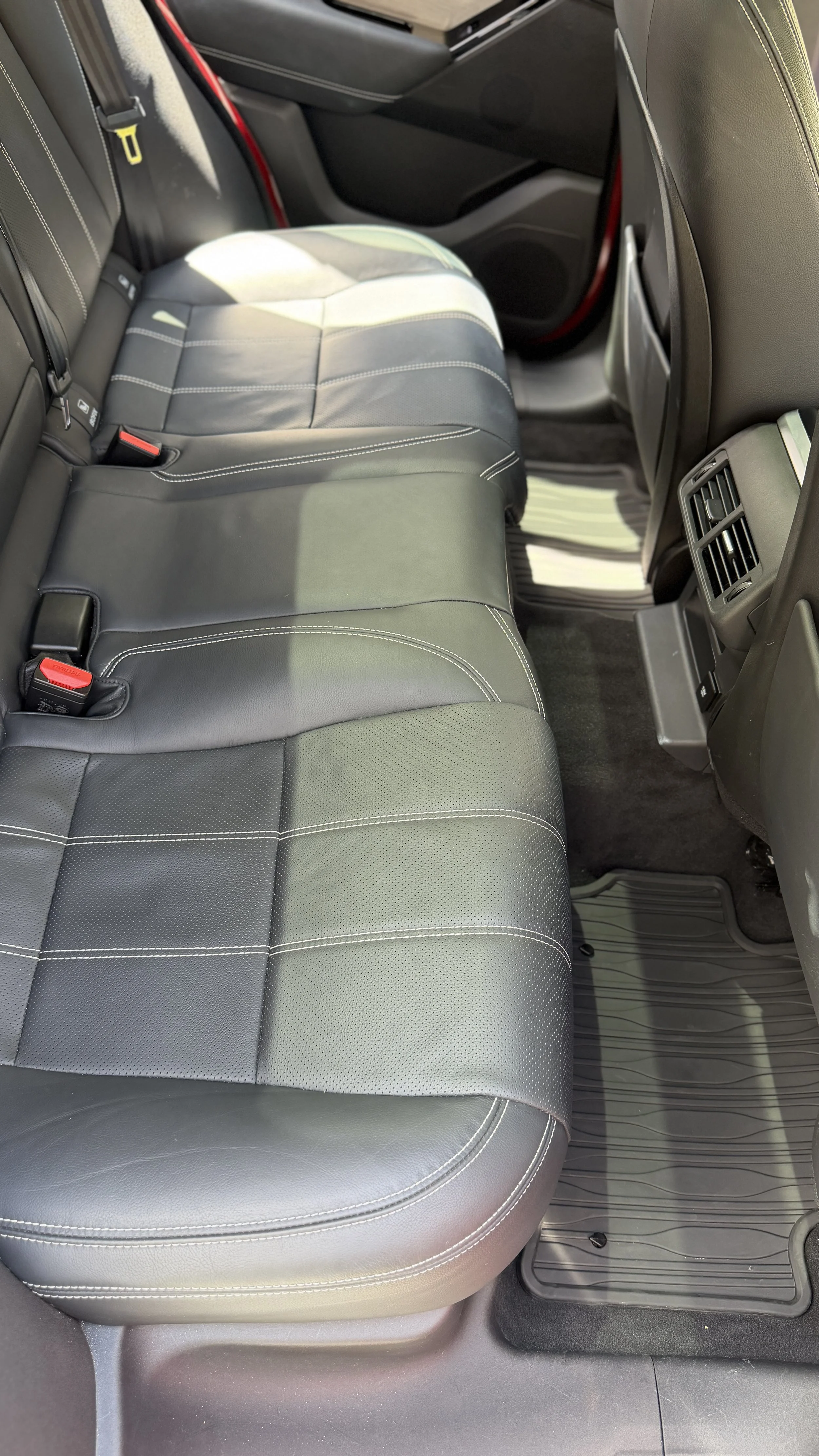 Backseat of a car with black leather seats, visible seat belts, car air vents, and gray floor mats.