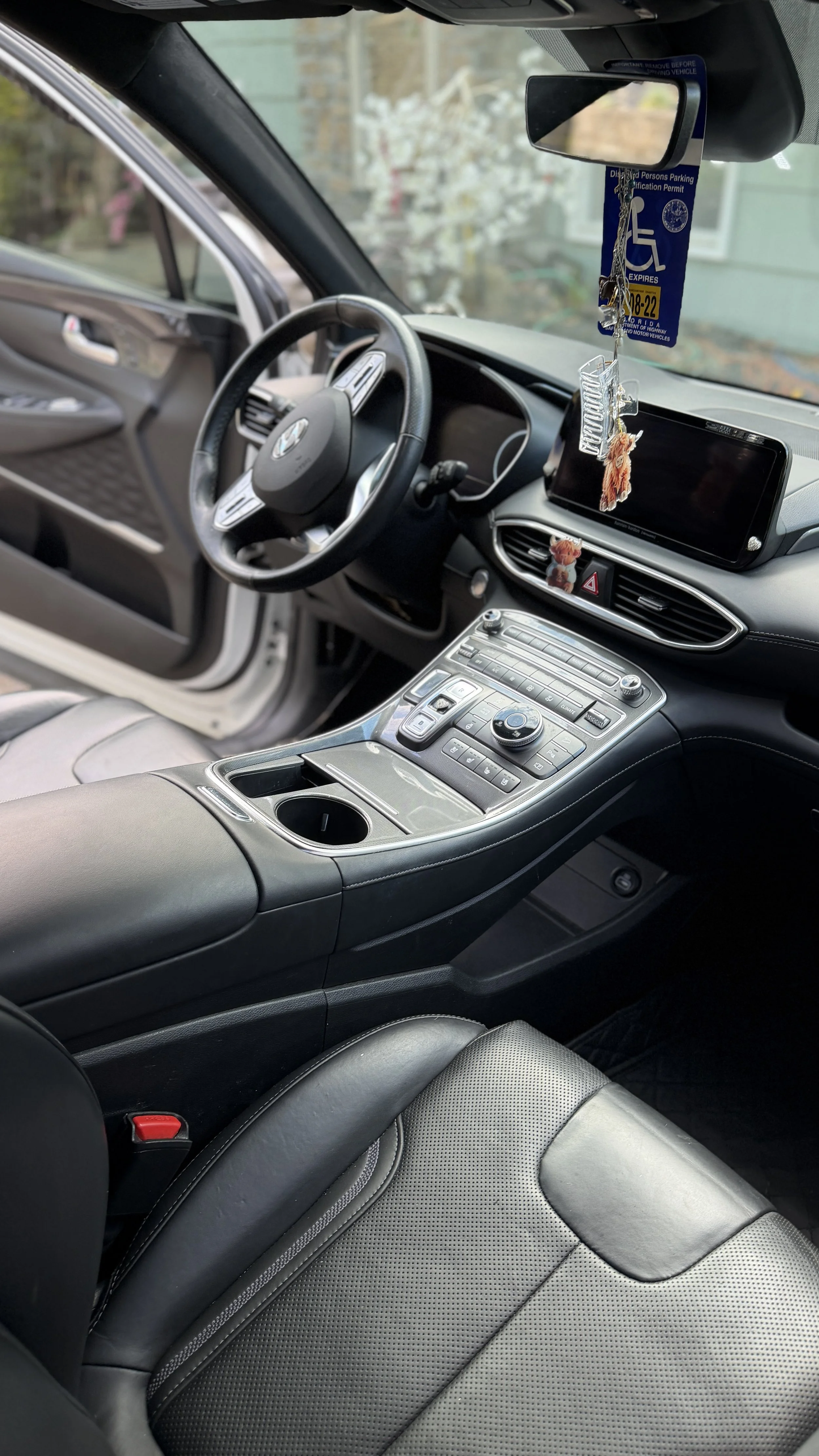 The interior of a black car showing the steering wheel, center console with controls, dashboard, and part of the driver's seat. The rearview mirror hangs with decorative charms and a parking permit. Visible outside are some trees and a house.