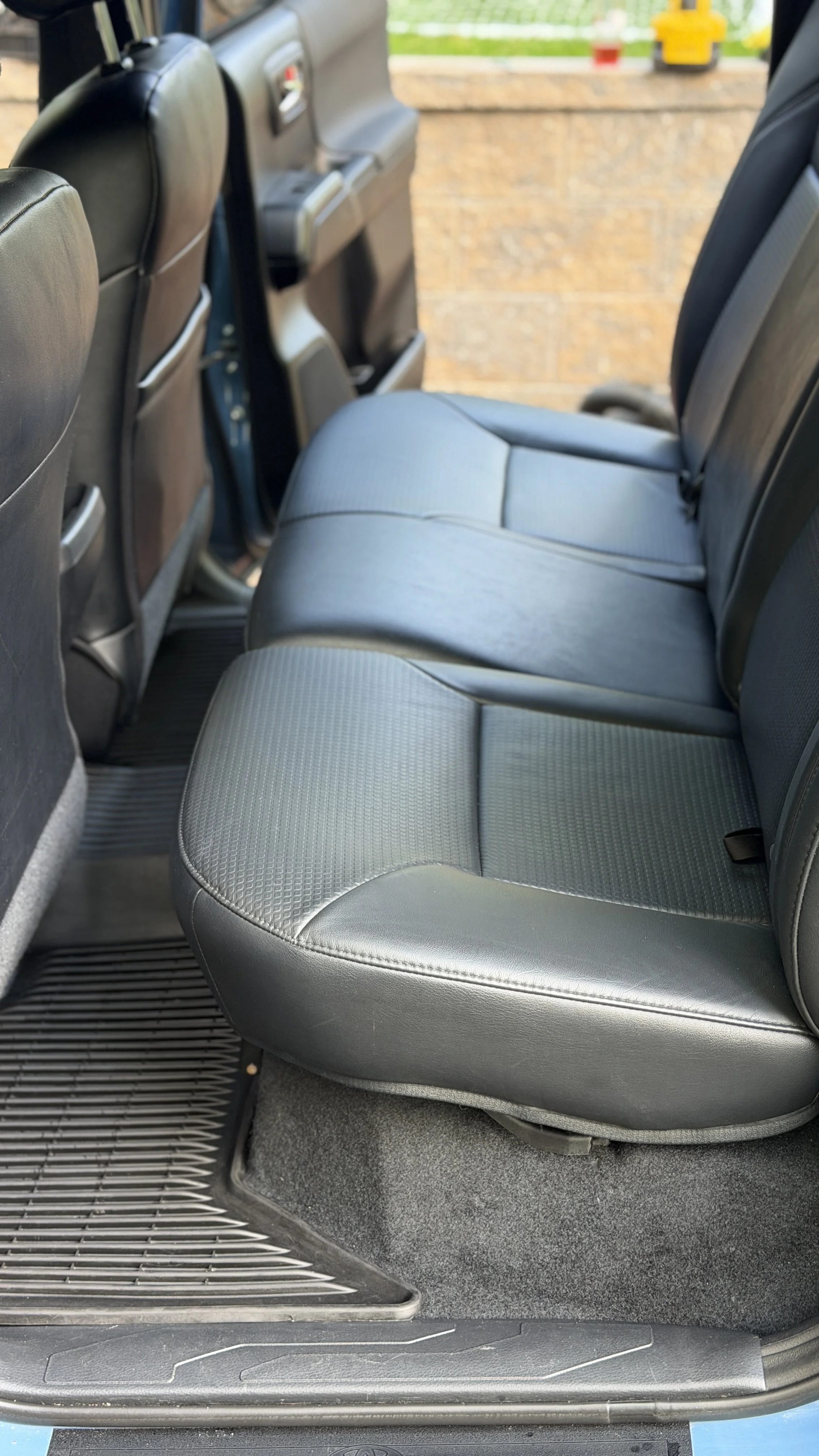 The backseat area of a vehicle showing gray fabric and leather seats, floor mats, and a view of a brick wall and outdoor scenery through the window.