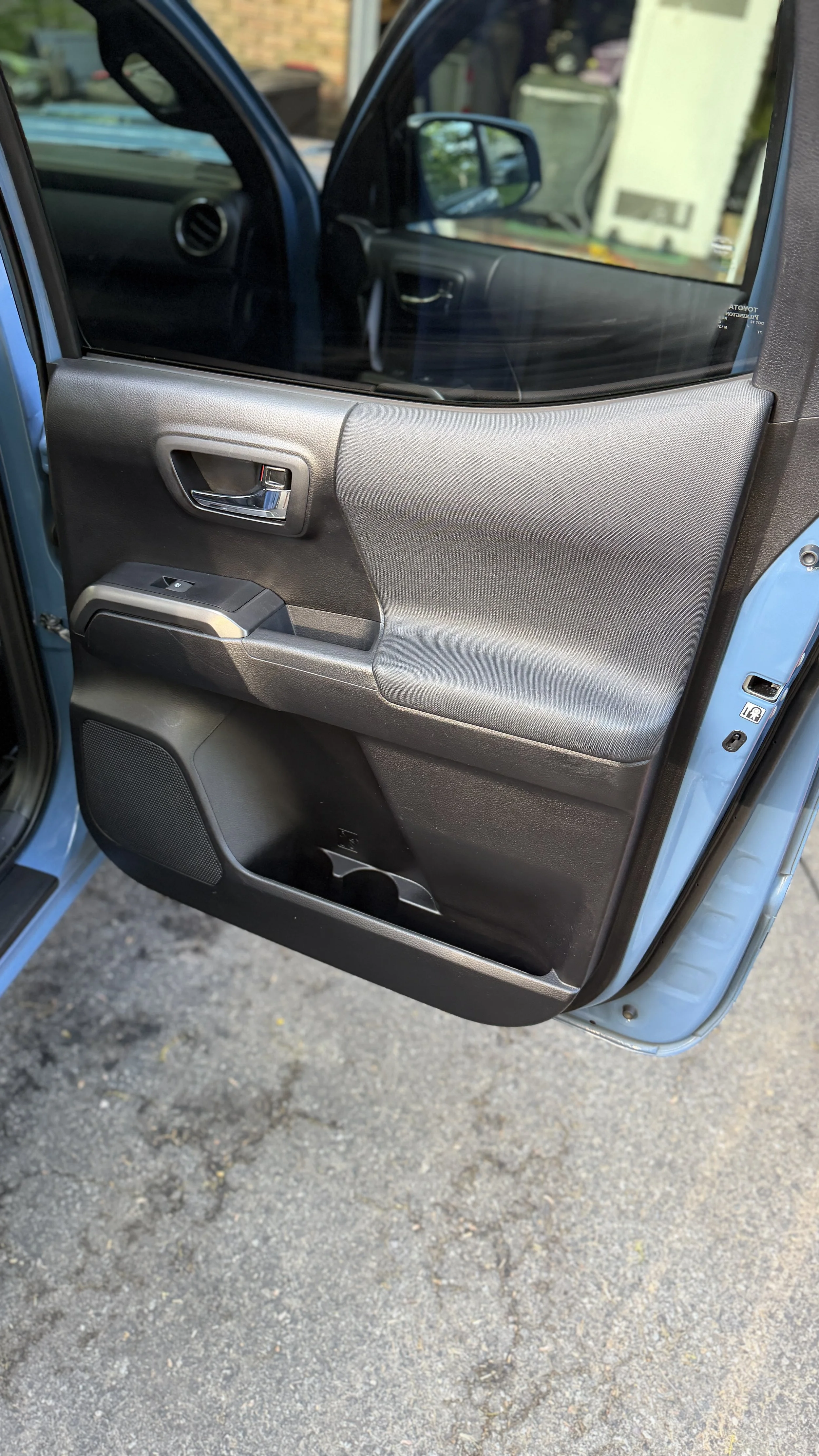 Close-up of the interior passenger side door of a blue vehicle, showing the door handle, window controls, and speaker, with a view of the parking lot ground outside.