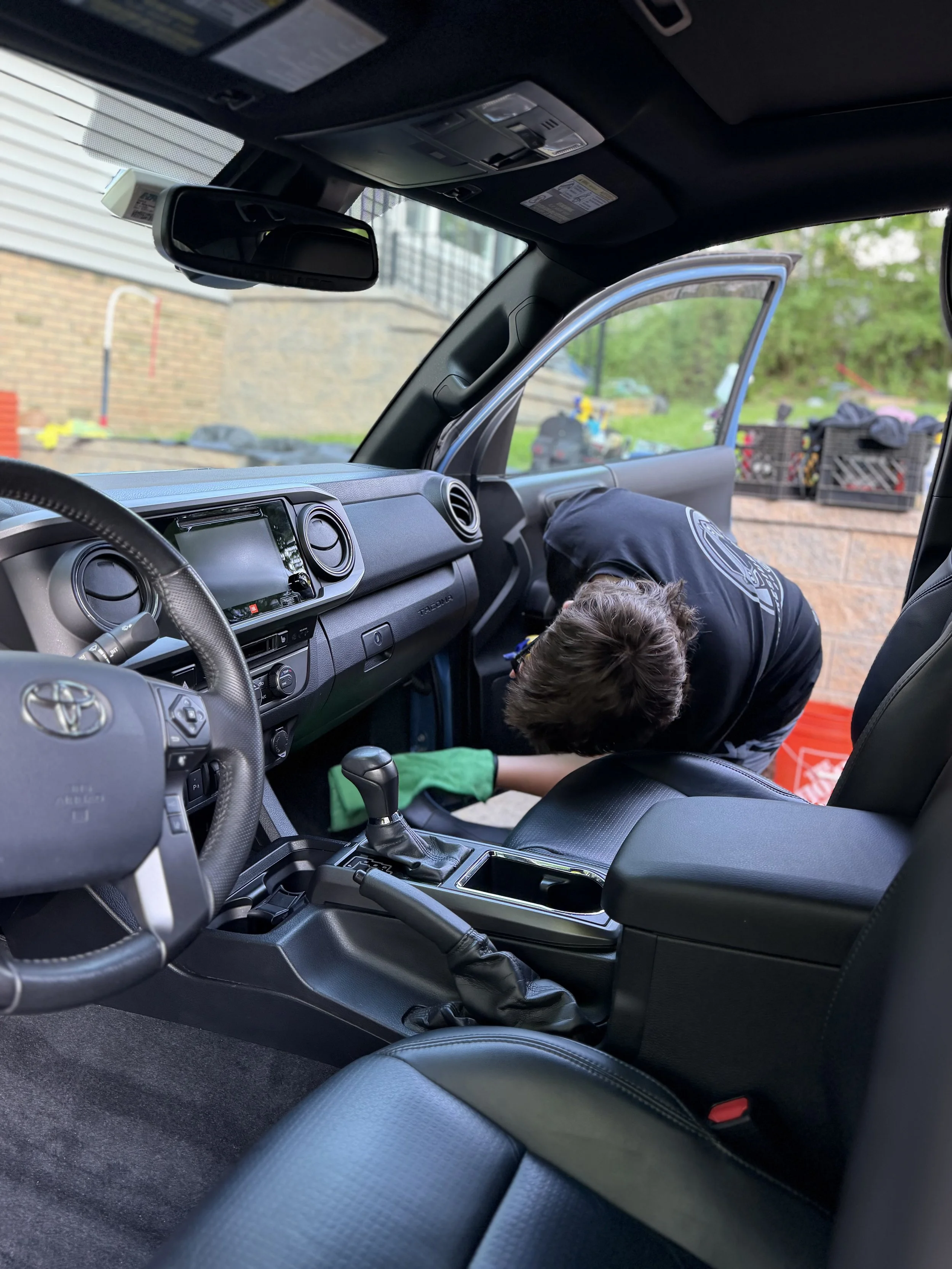 Inside a vehicle, a person is working near the passenger seat with the door open, outside are crates and items on the ground, and a brick building and greenery are in the background.