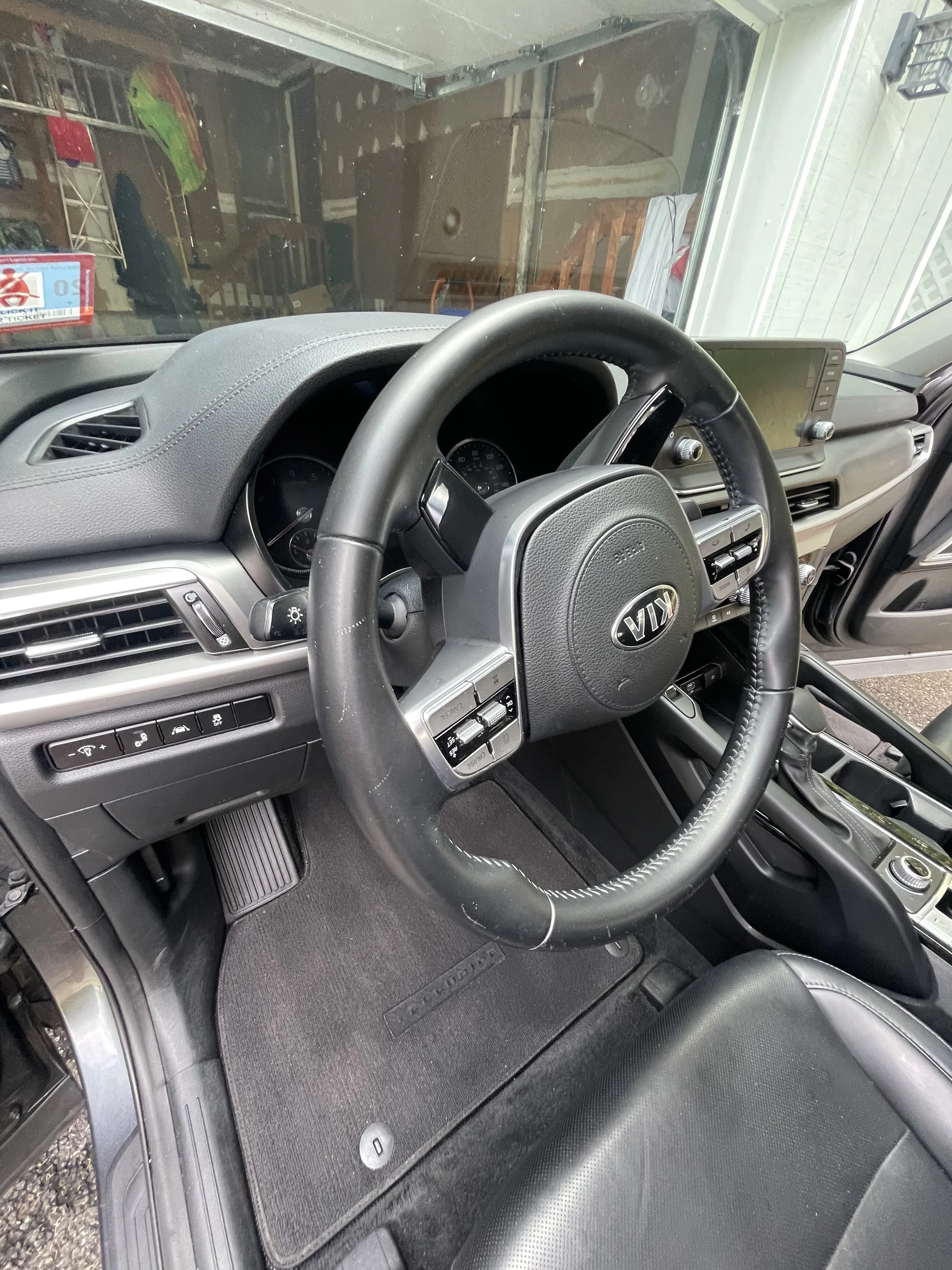 View from the driver's seat of a Kia vehicle showing the steering wheel, dashboard, and front interior. Through the windshield, a garage or storage area with various objects and wooden furniture is visible.