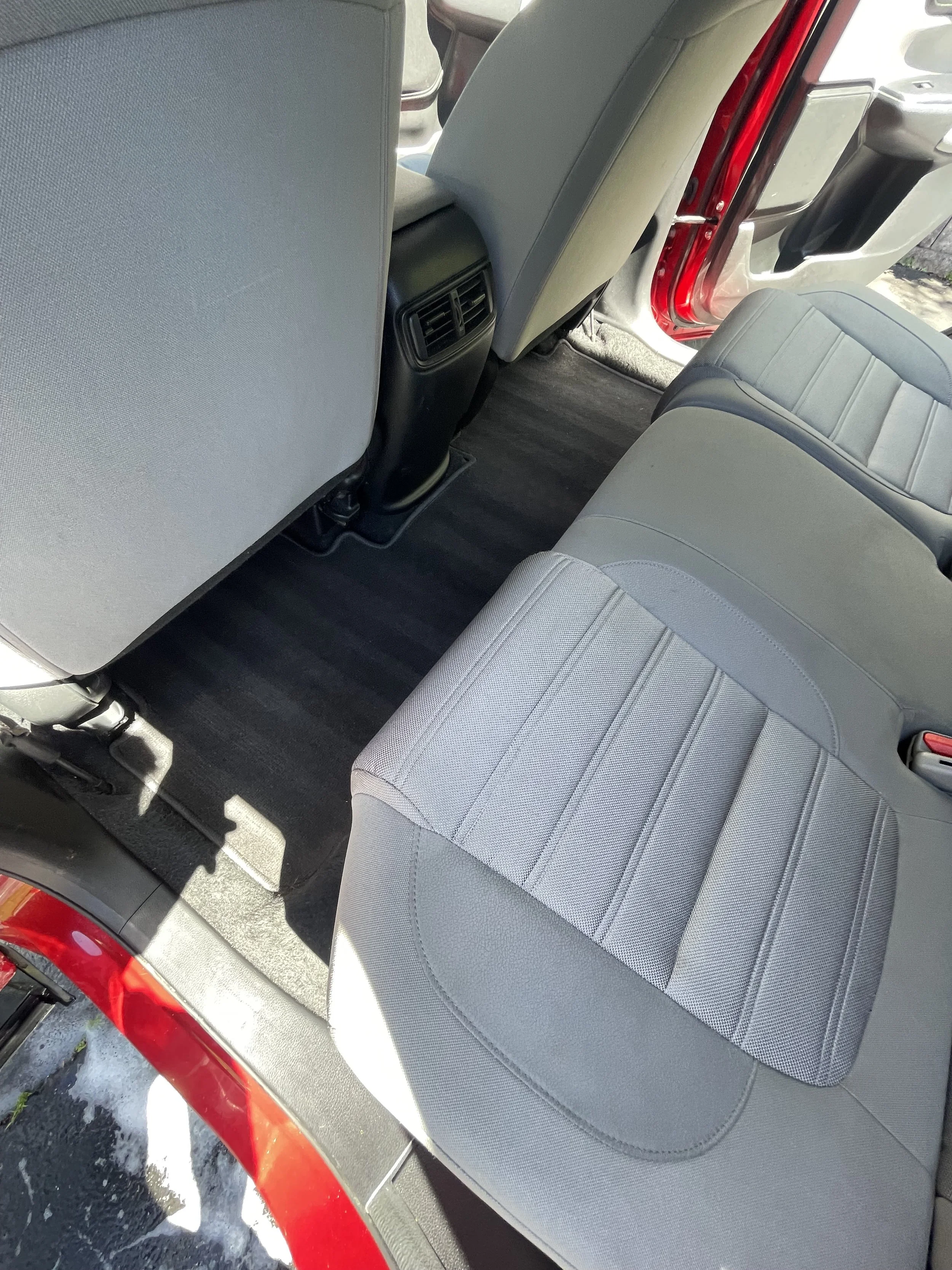 Interior of a car showing the back of the front seats, the rear seats, and part of the door frame. The seats are gray with a textured fabric pattern, and the flooring is dark. There is also an air vent on the back of the center console.
