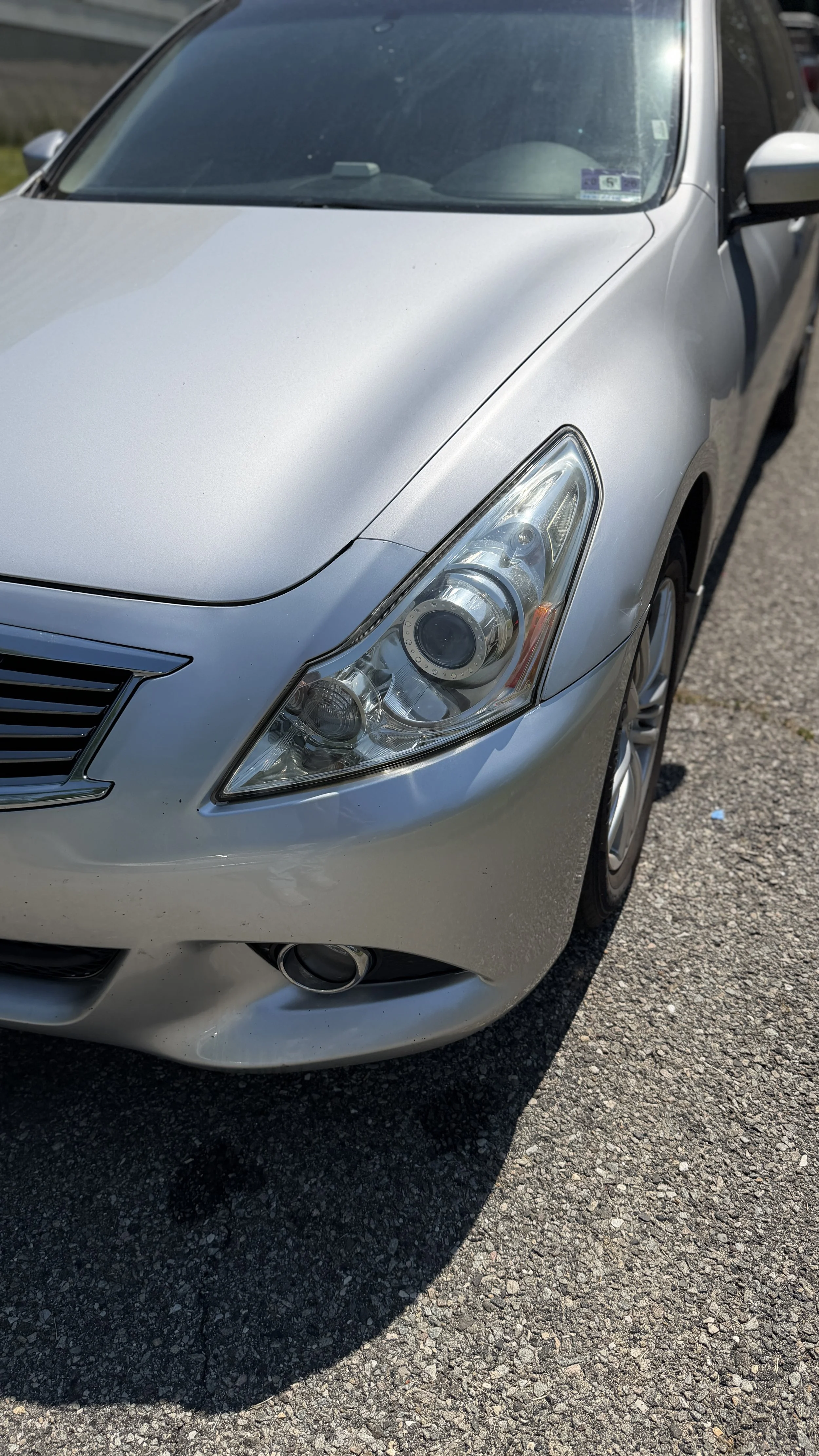 Close-up of a silver car's front right side, showing the headlight, grille, and part of the windshield, parked on an asphalt surface.