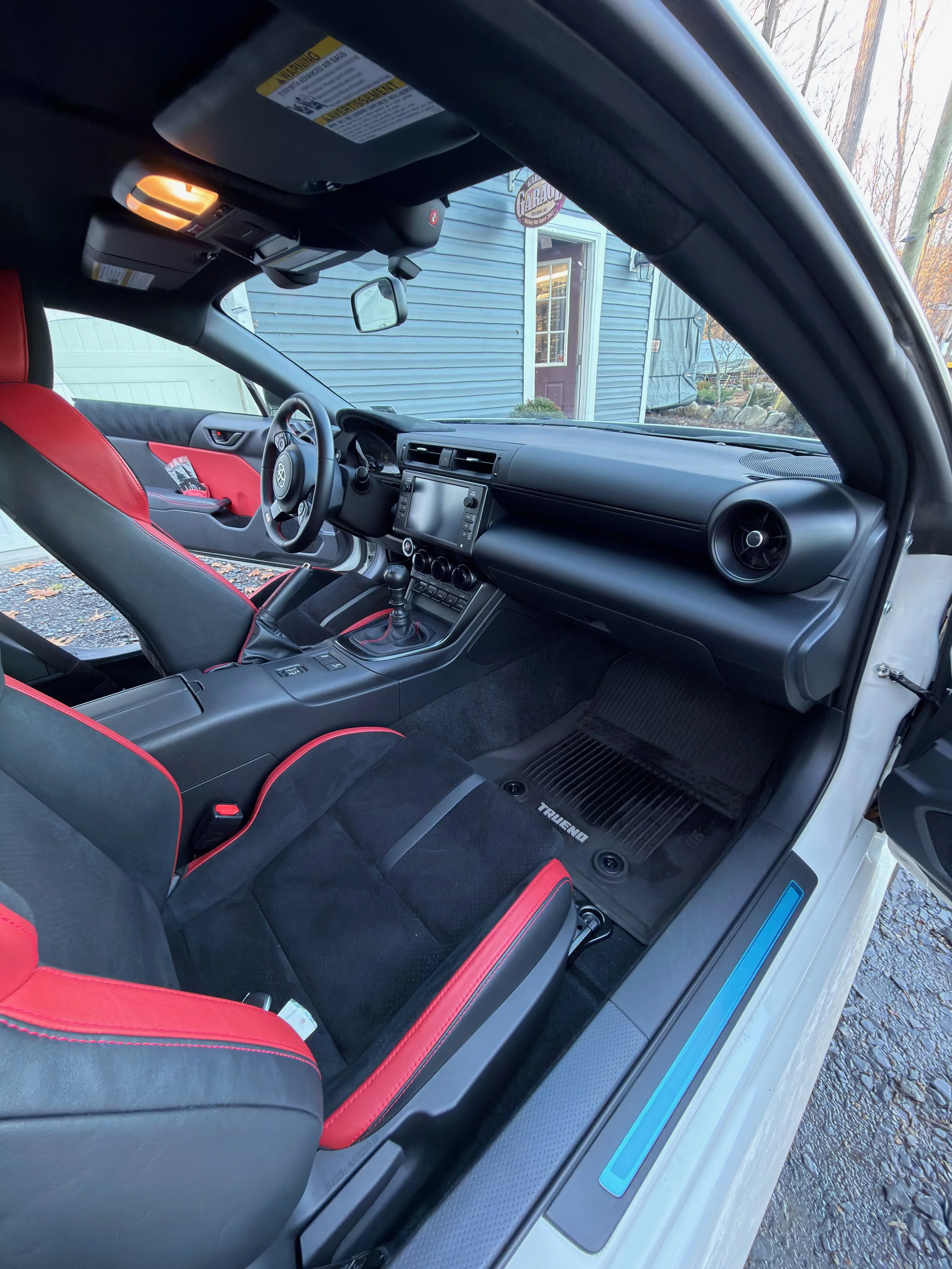 The interior of a car showing the driver's side with a black dashboard, leather seats with red accents, a gear shift, and a touchscreen display. The view through the windshield shows a blue house with a sign and some trees outside.