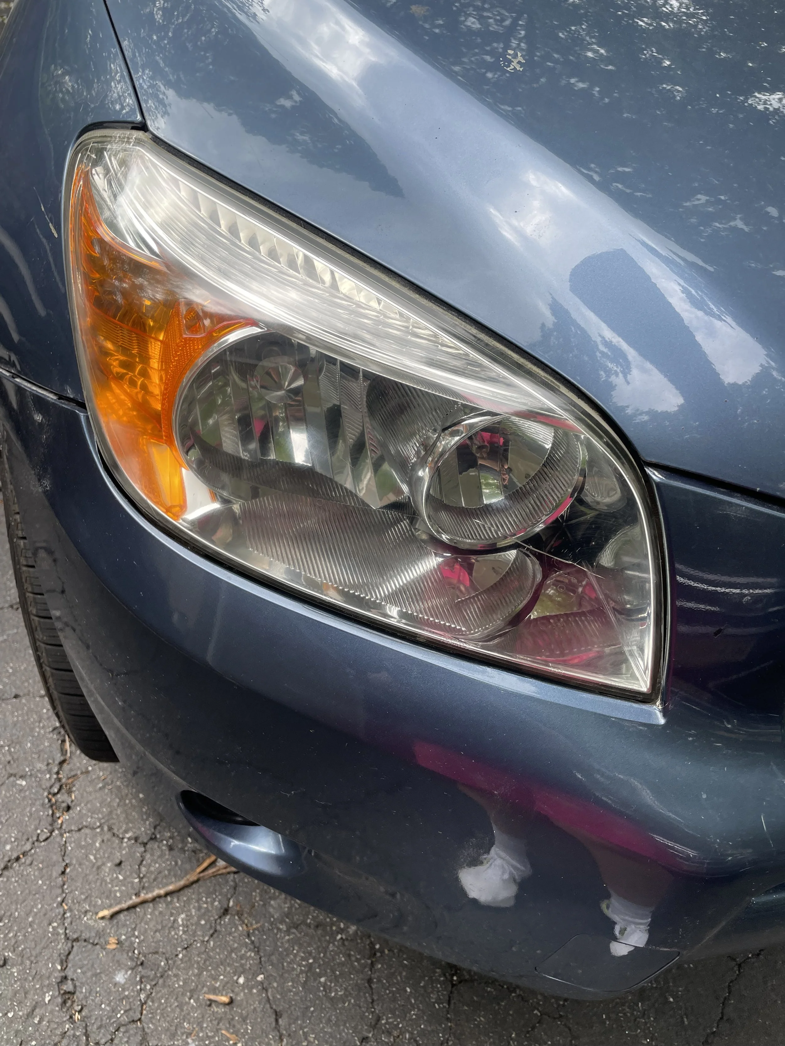 Close-up of a blue car's headlight and front bumper with reflected trees and sky, parked on rough asphalt with small cracks.