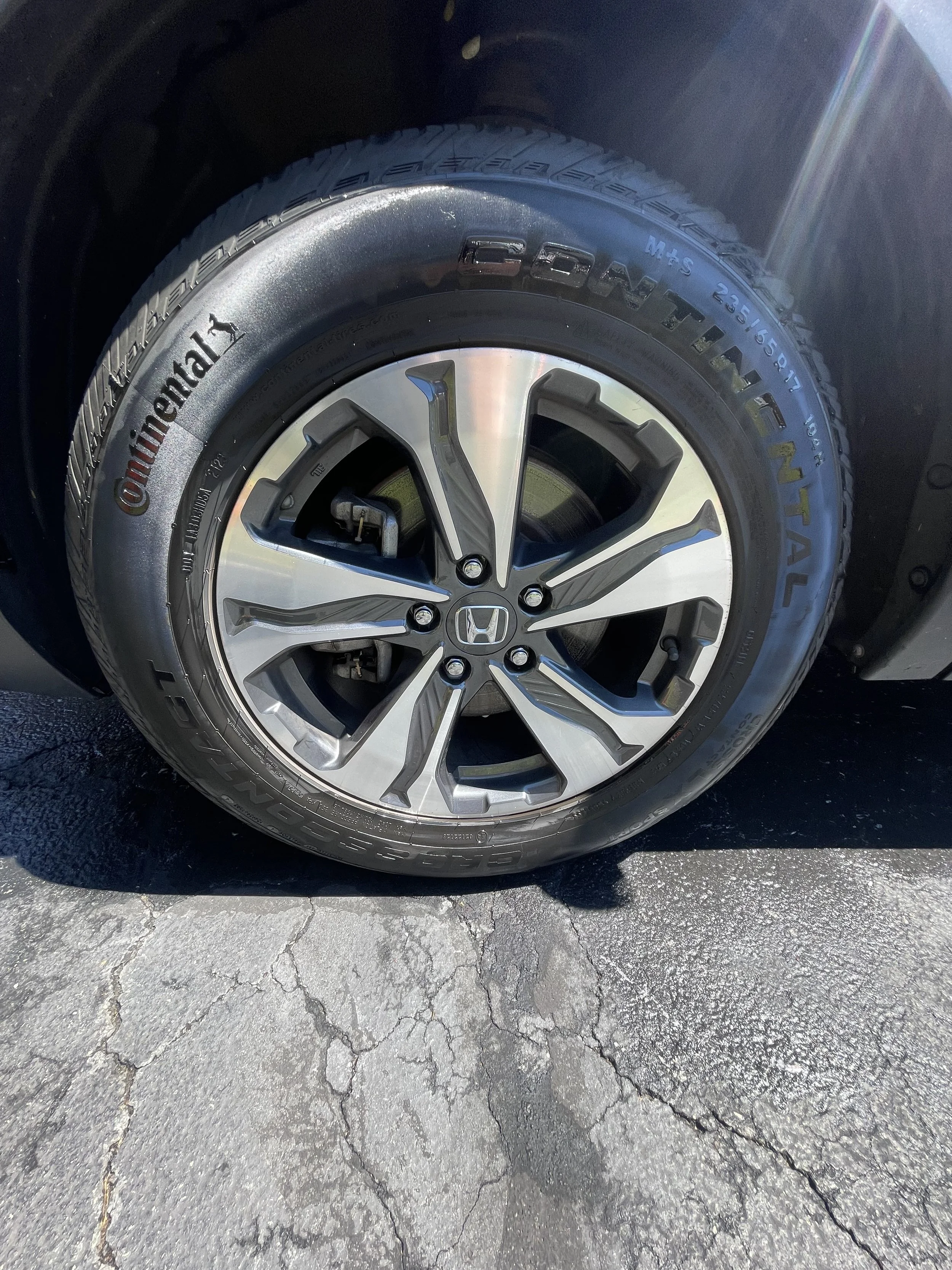 A close-up of a car tire with a silver alloy wheel. The tire brand is Continental, and the rim features a Honda logo. The car is parked on a cracked asphalt surface with sunlight casting a reflection on the tire.