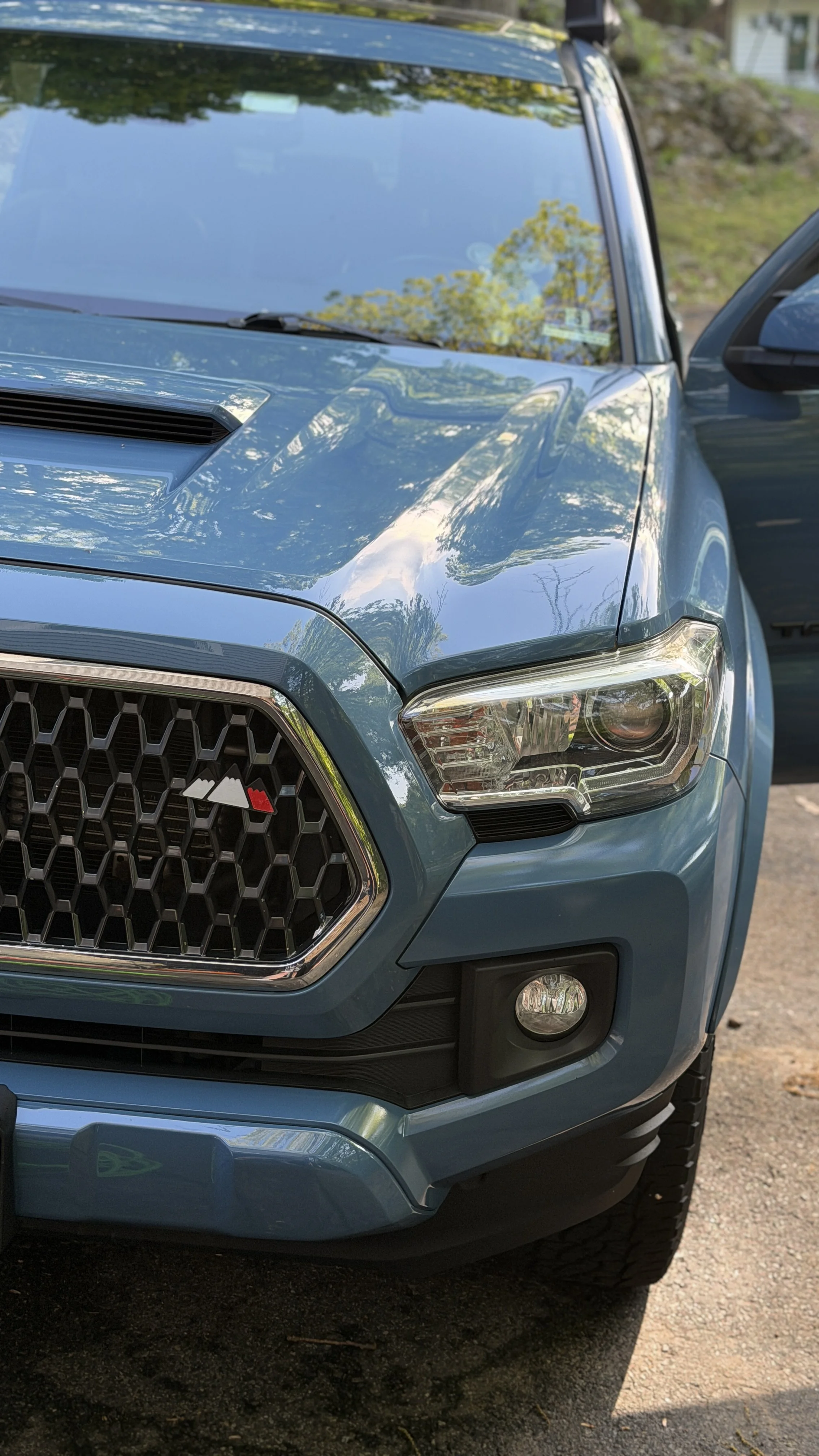 Close-up of a blue pickup truck's front corner, showing the grille, headlight, and fog light, with reflections of trees on the windshield.