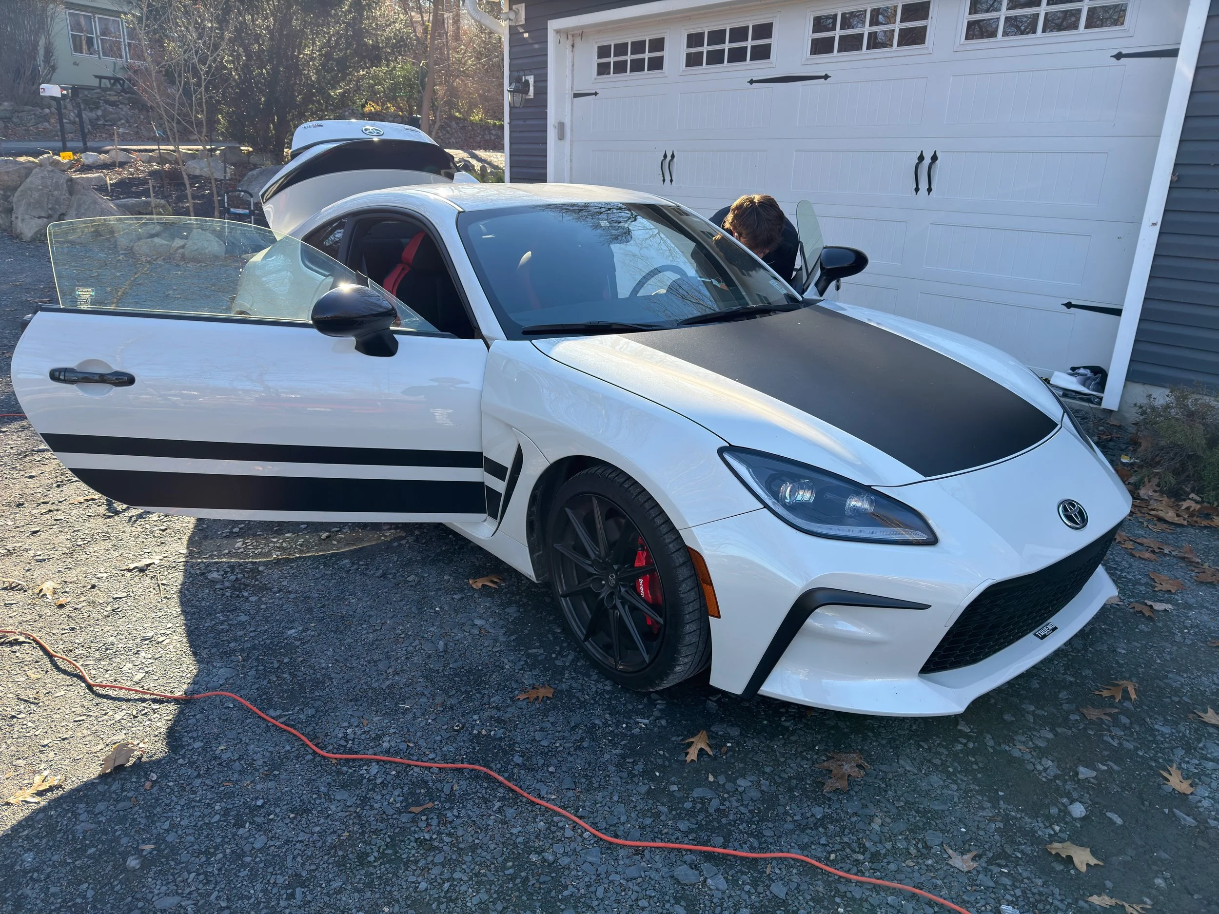 A white sports car with black accents and red interior is parked in a driveway with the driver's side door open. A person is leaning into the car near the front passenger seat. The background includes a gray garage door, trees, large rocks, and a basketball hoop.