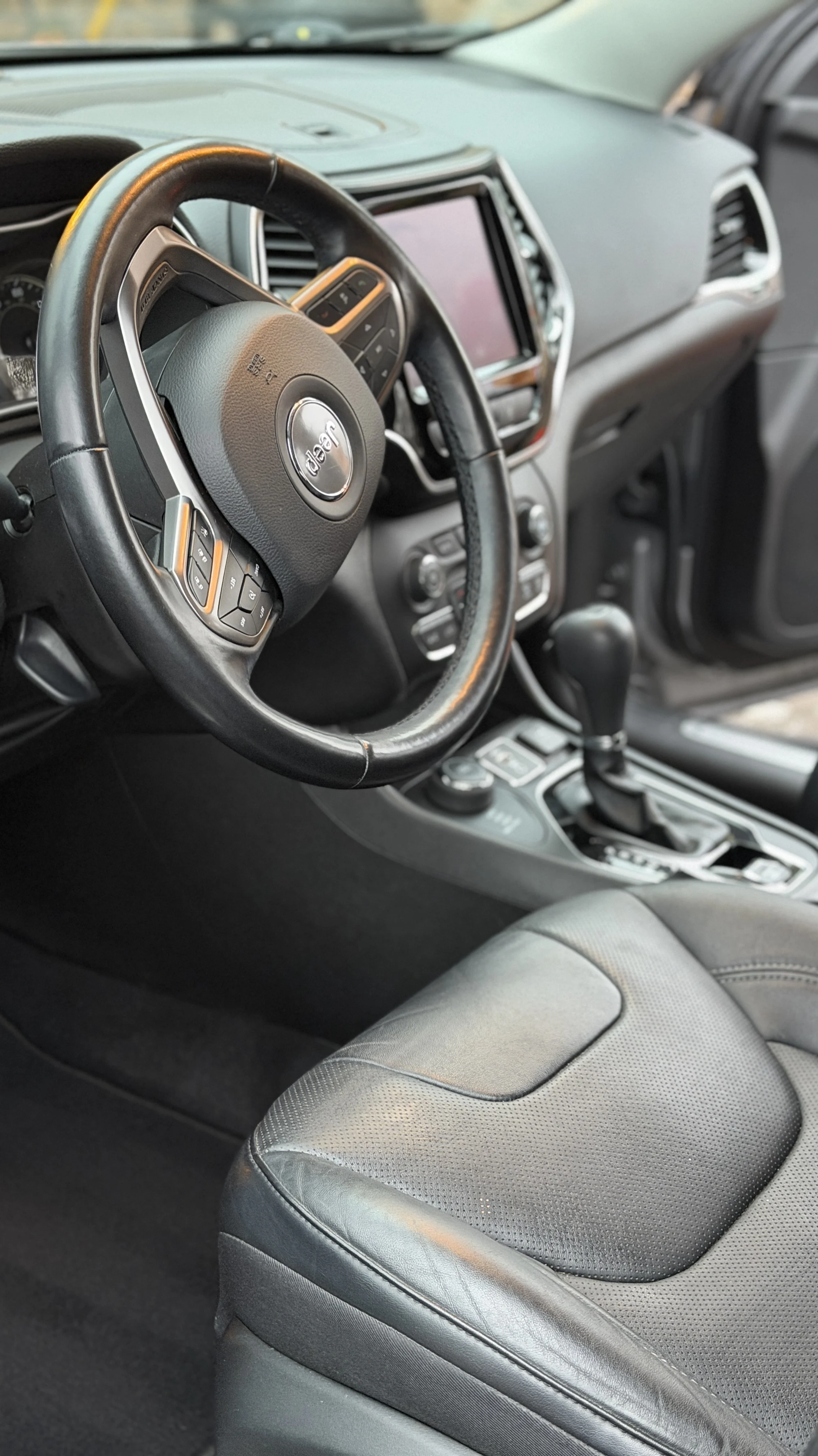 Inside view of a car dashboard and front passenger seat, featuring a steering wheel with Jeep logo, gear shift, and infotainment screen.