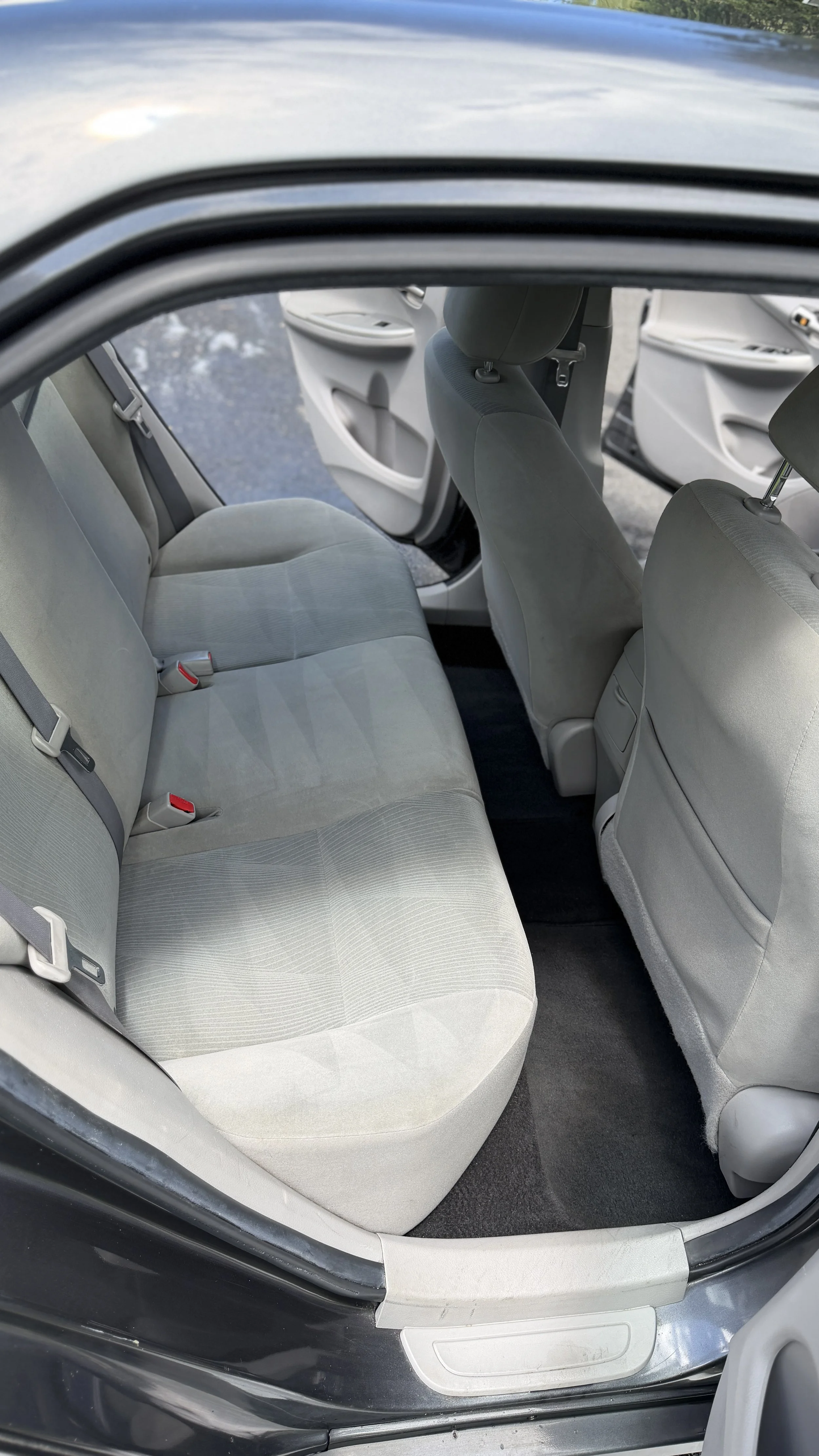 Photo of the interior of a car showing the back seat and part of the front seats, with gray fabric upholstery, seat belts, and a black carpeted floor.
