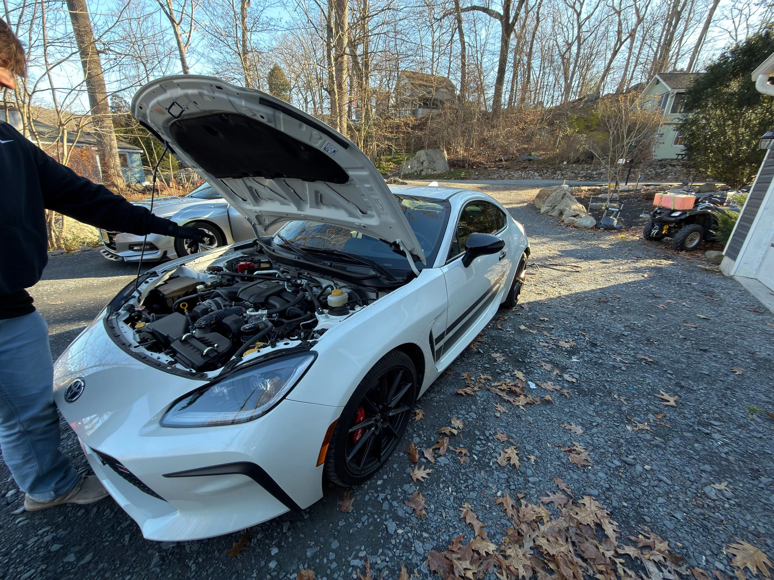 A person checking the engine of a white sports car with its hood open in a driveway surrounded by trees with fallen leaves, houses, and other parked vehicles in the background.