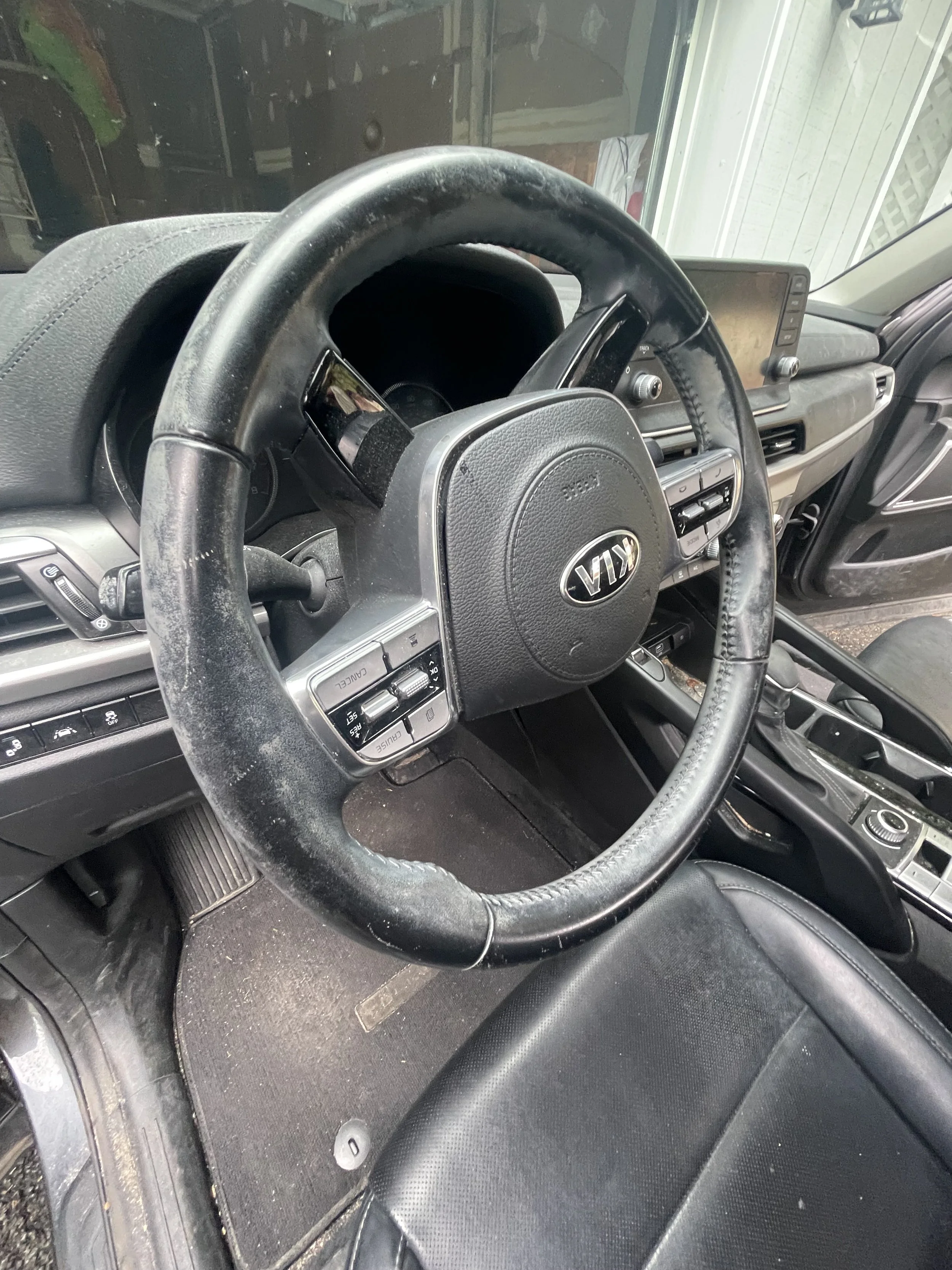 Interior view of a vehicle showing a steering wheel, dashboard with controls, and a digital display screen. The dashboard and steering wheel are dusty, and the flooring appears dusty and dirty.