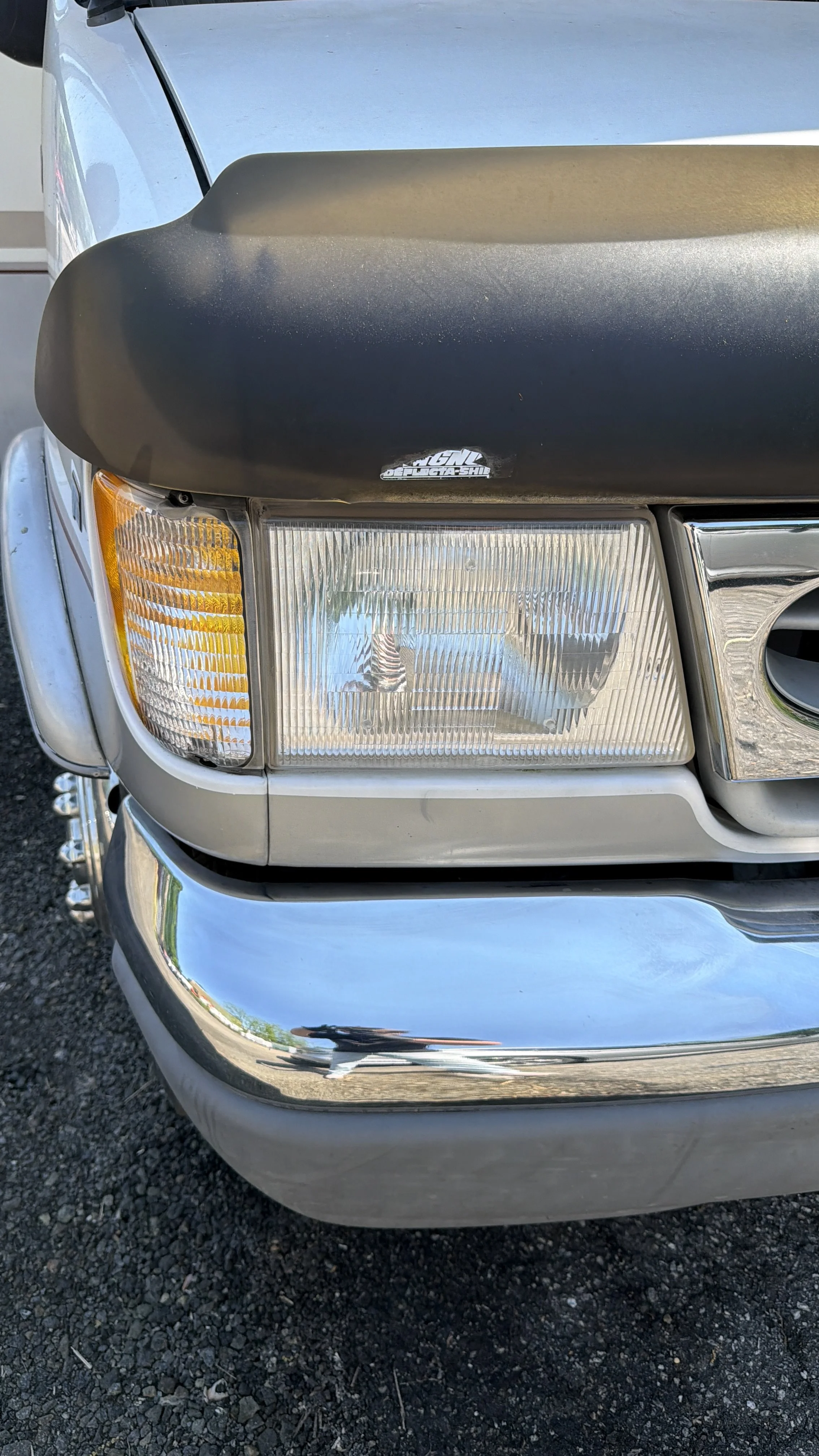 Close-up of the front of a silver vehicle, showing the headlight, grille, and a black hood ornament.