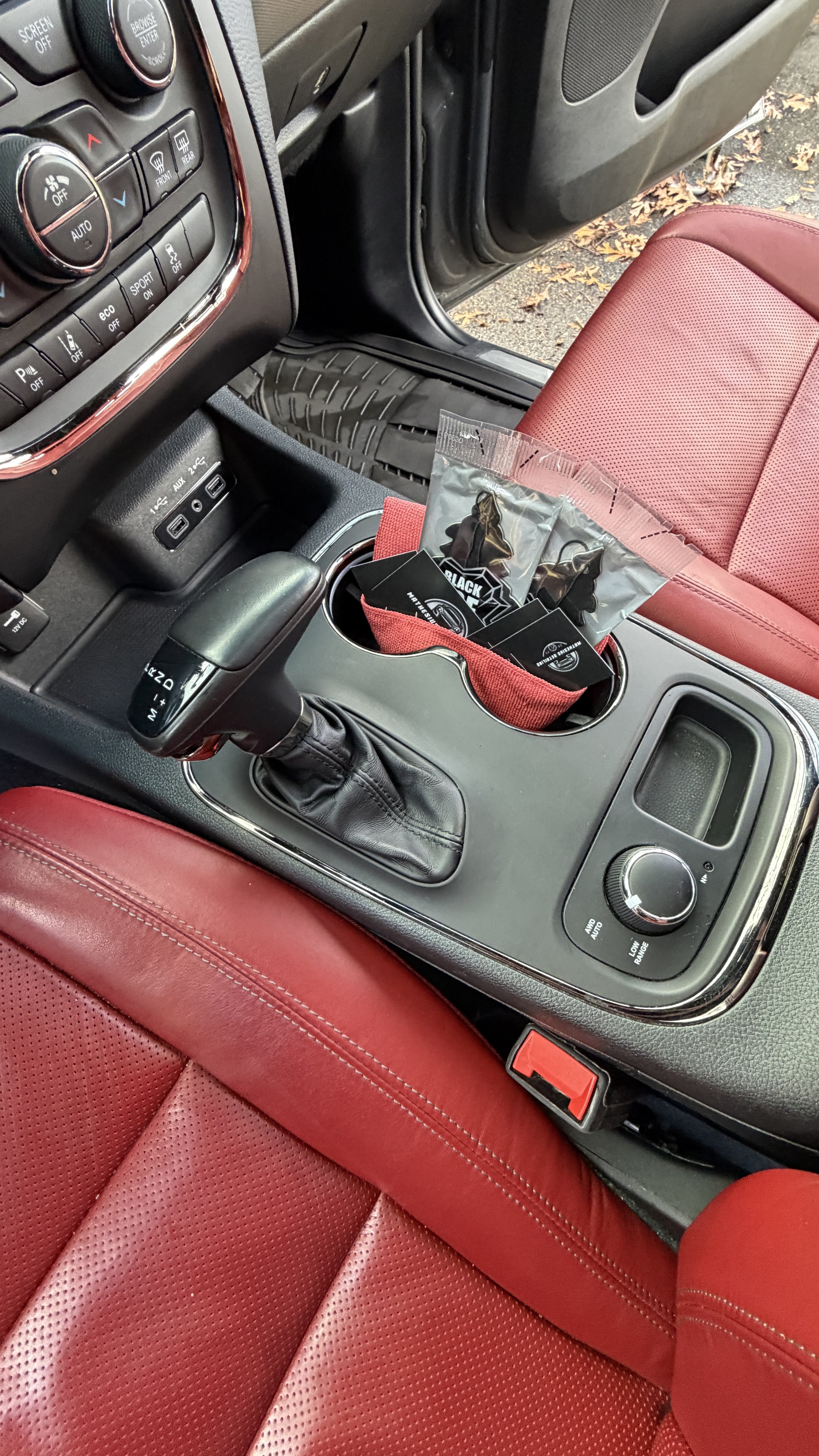 Inside a vehicle, showing the center console with gear shift, cup holder with rubber gloves and a small bag, red leather seats, and part of the dashboard with climate controls.