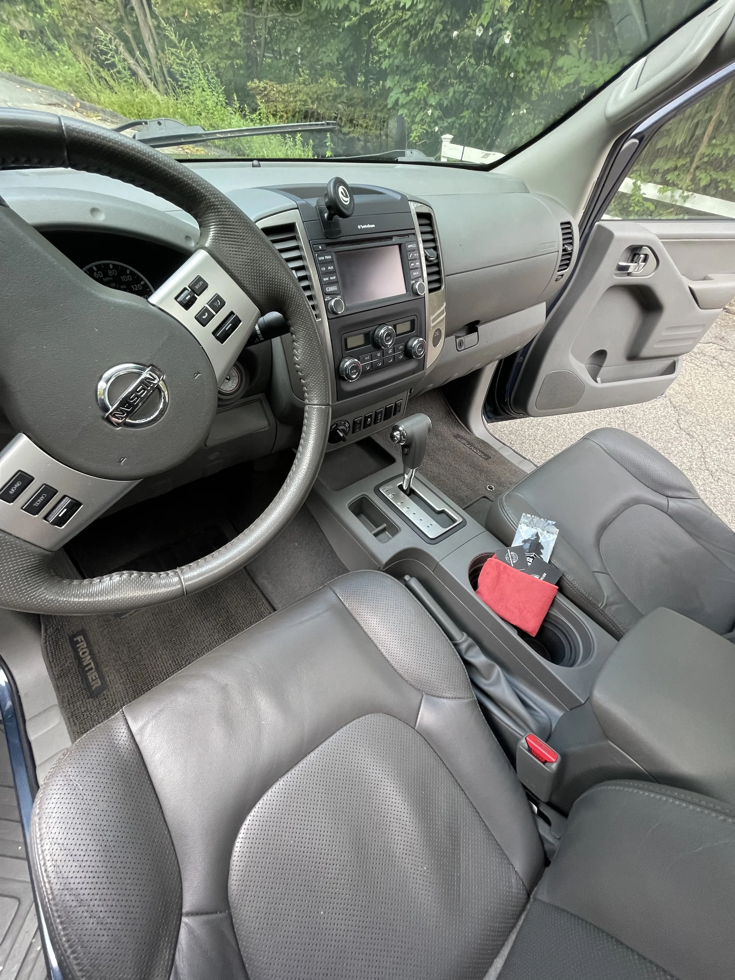 Interior of a Nissan vehicle showing the steering wheel, dashboard, center console with gear shift, and front passenger seat with a red cloth and a small packet on it.