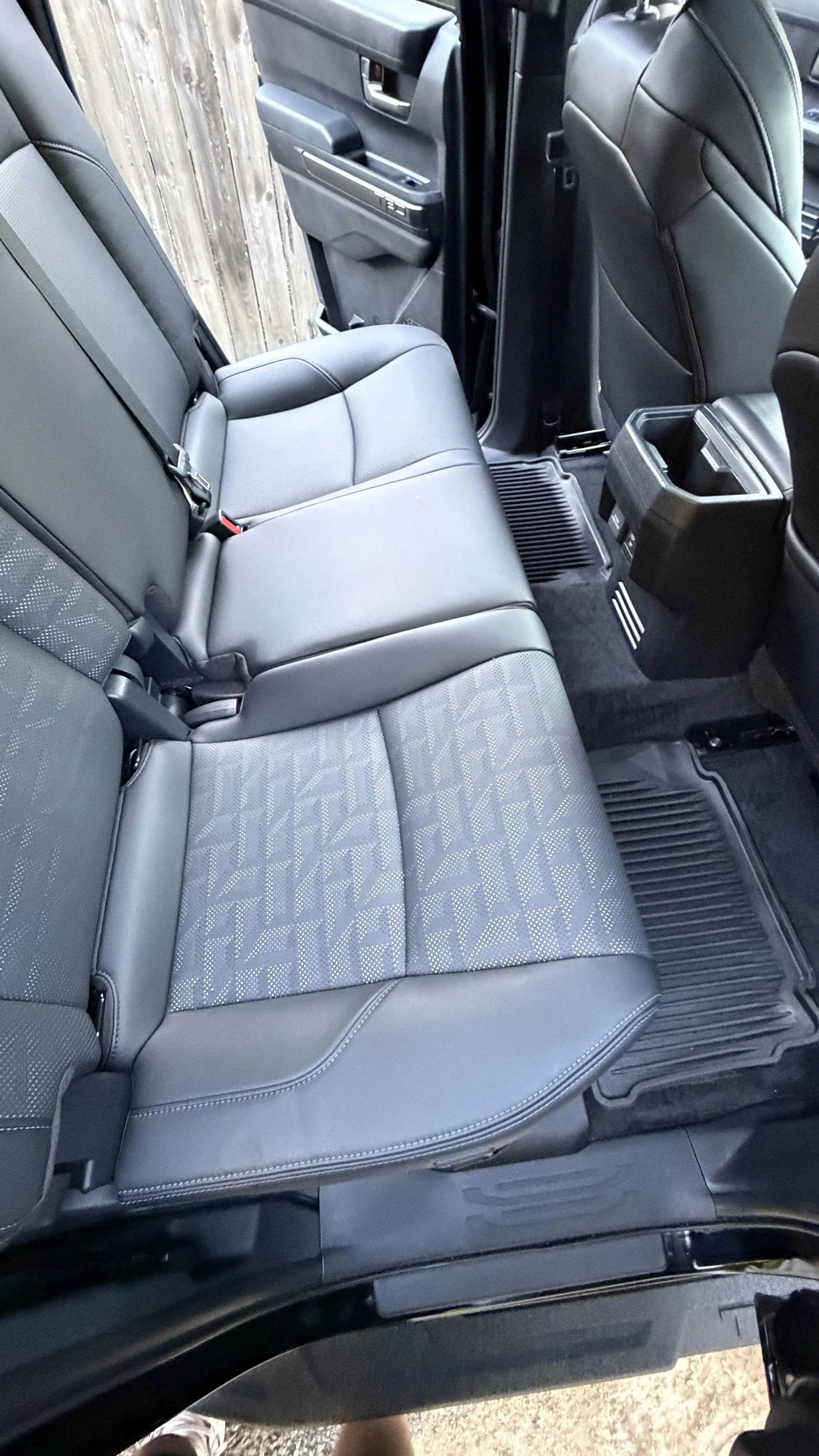 The interior of a vehicle showing the back seat, with the left side partially open door, black leather seats, and a fence outside.