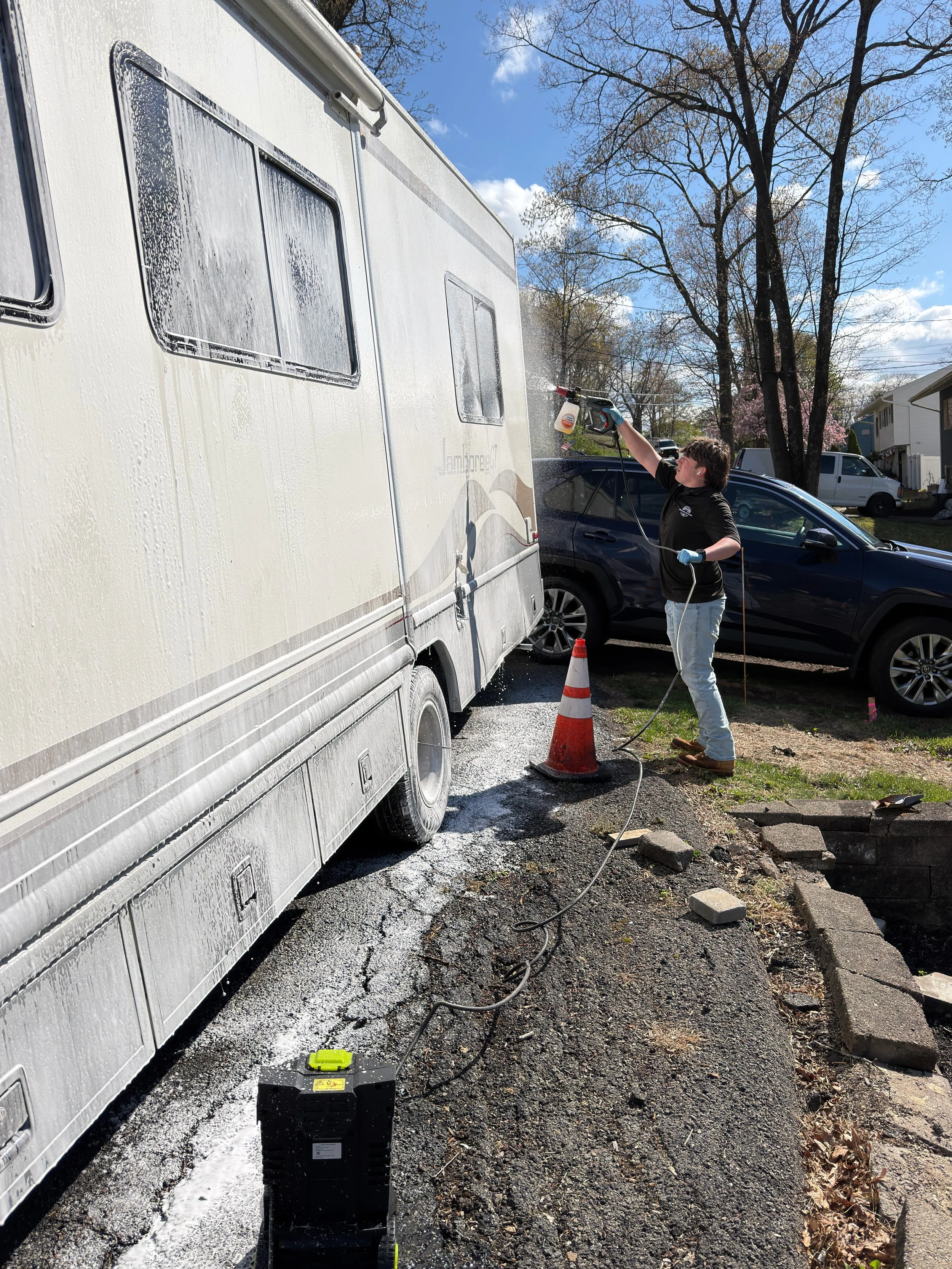 A person pressure washing the side of a white camper trailer, with soap and water streaming down, on a paved driveway with orange cones and a black toolbox nearby.