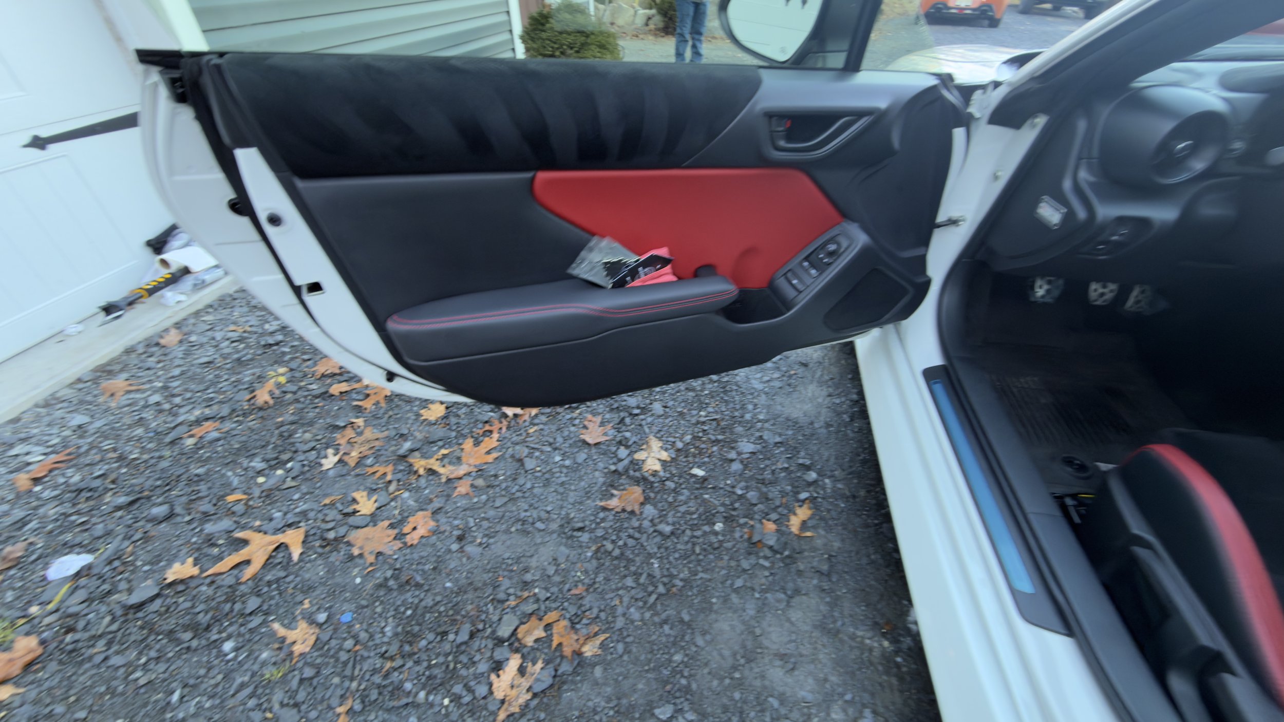 The interior of a car showing the driver's side door and part of the dashboard. The door has black and red interior panels with window controls. The car is parked outside with fallen leaves on the ground.