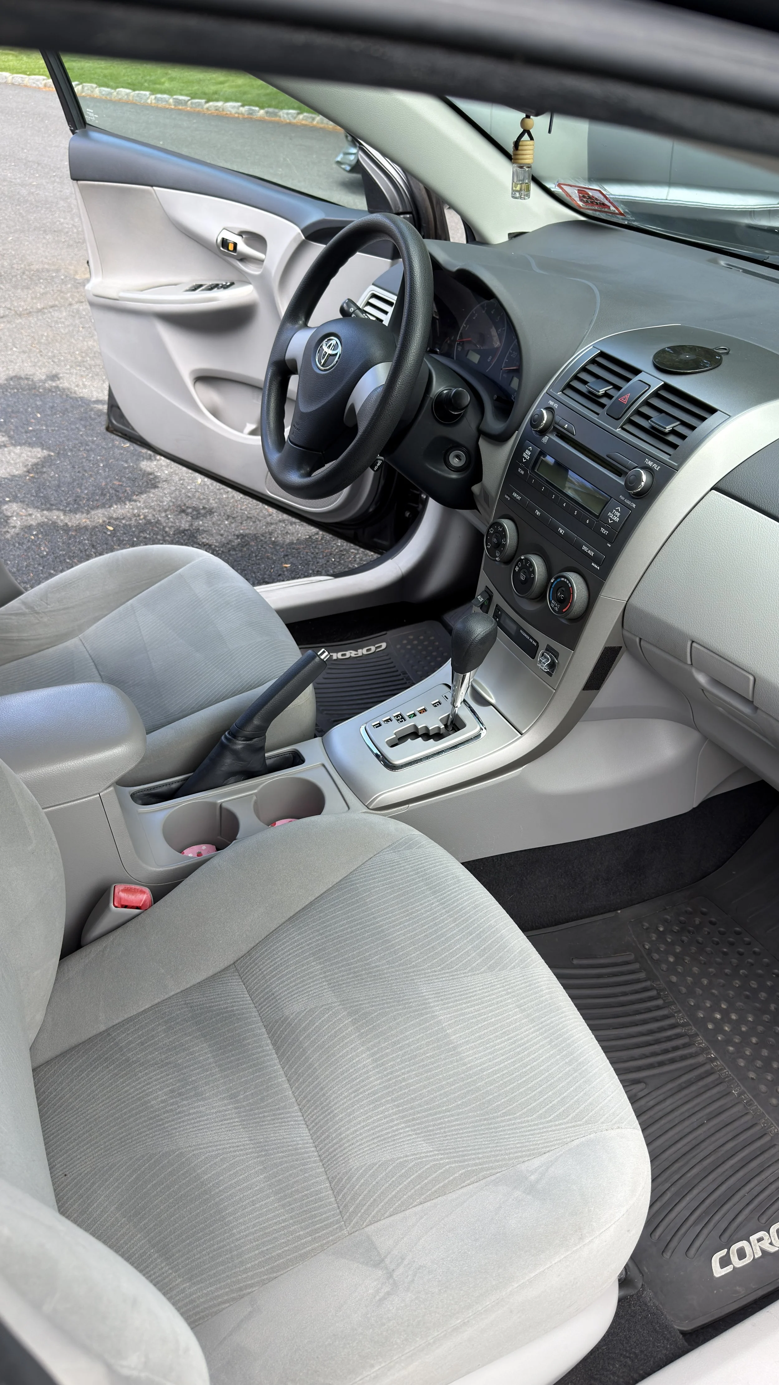Interior of a light gray Toyota Corolla with a manual transmission, front driver seat, dashboard, steering wheel, and open driver's side door.