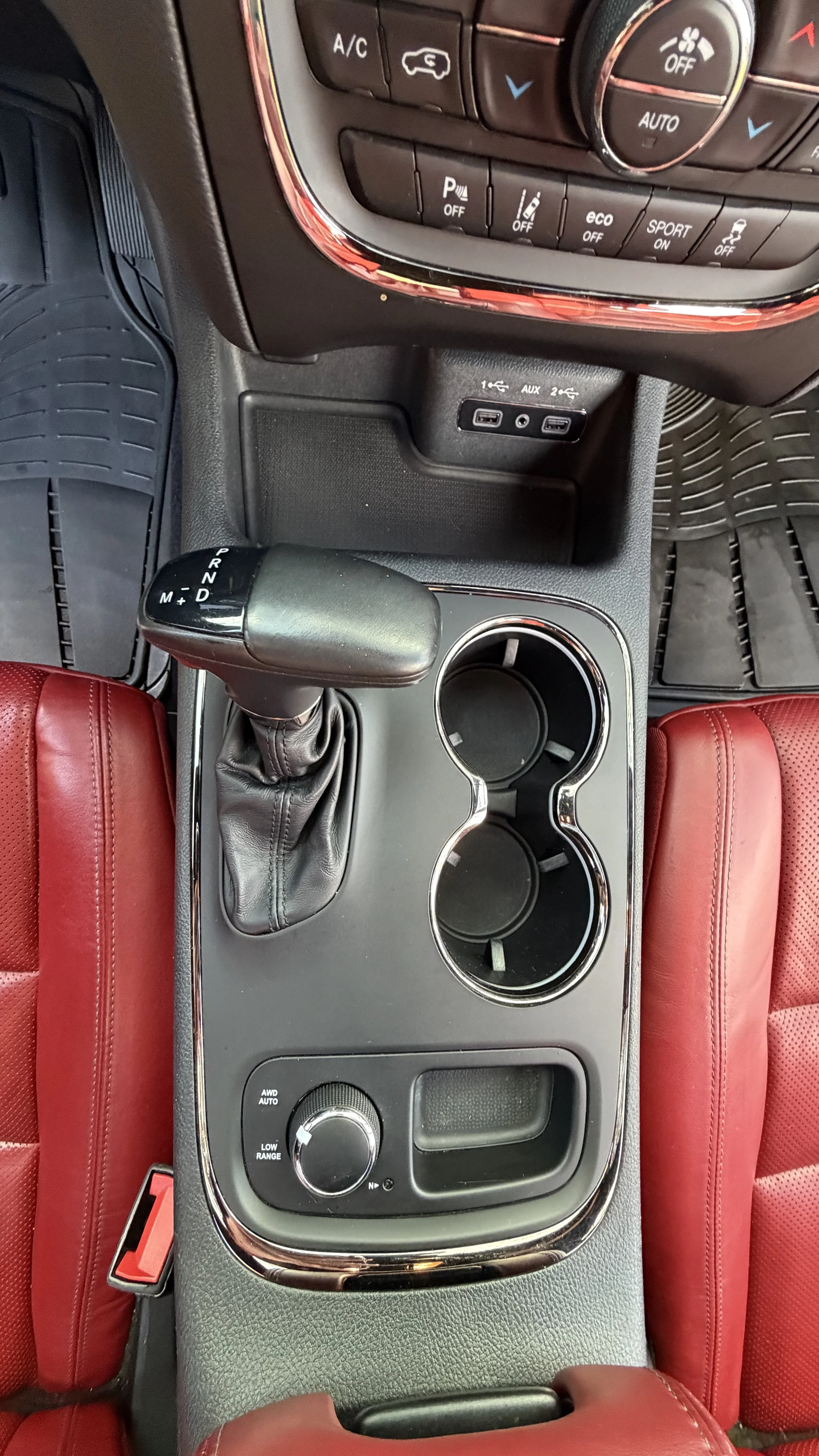 Center console of a vehicle with gear shift, cup holders, and control buttons, surrounded by red leather seats.