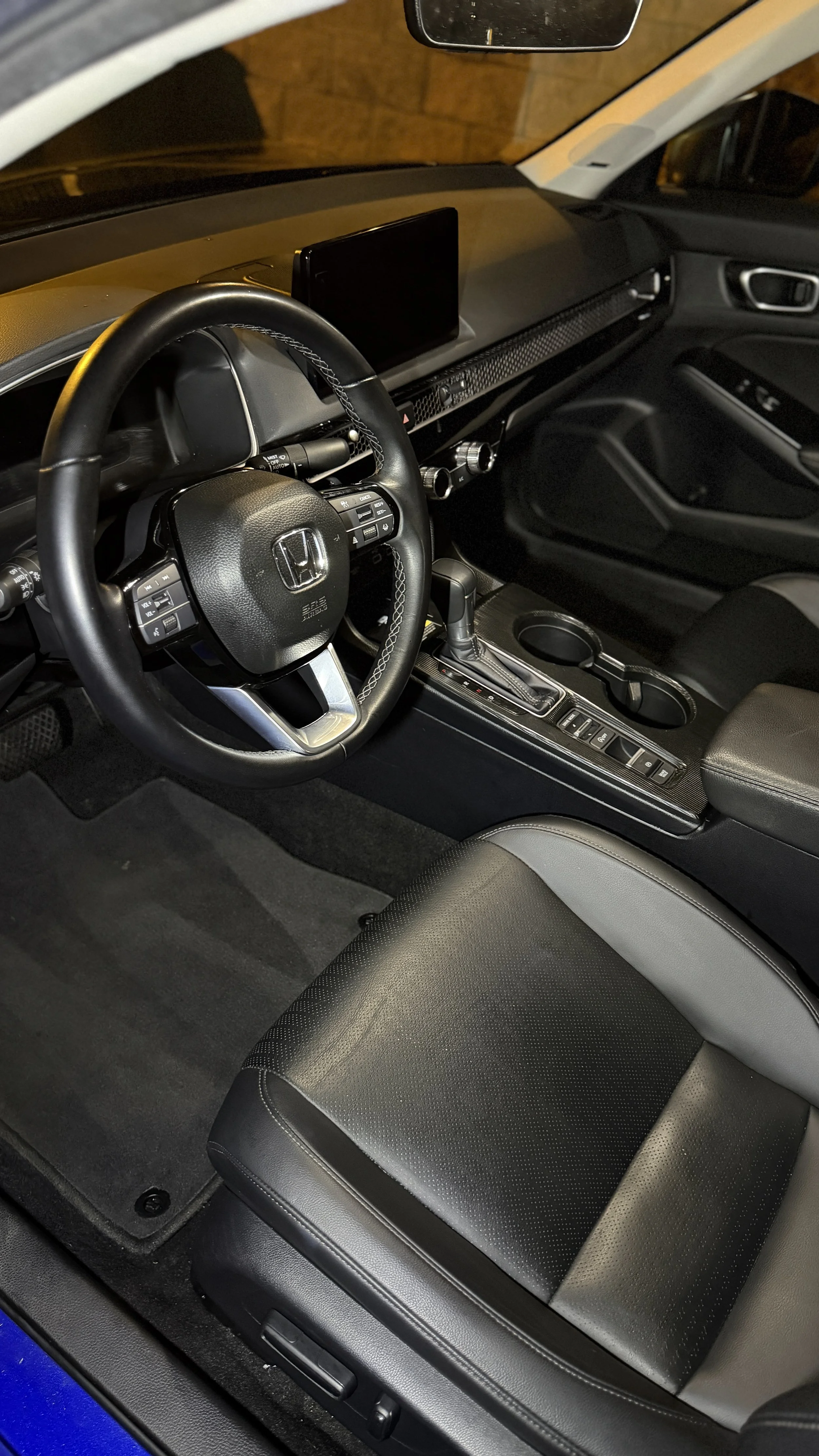 Interior of a Honda vehicle showing the steering wheel, dashboard with a digital display, gear shift, cup holders, and the front seats, with the steering wheel featuring Honda's logo.