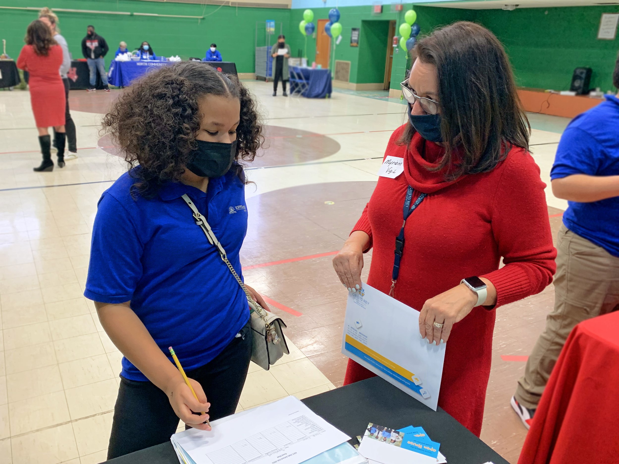 A young girl and a woman wearing masks at an event, signing a document. The woman wears a red sweater and glasses, holding a large paper, and the girl is in a blue jacket with a lanyard, holding a pencil and writing on paper.