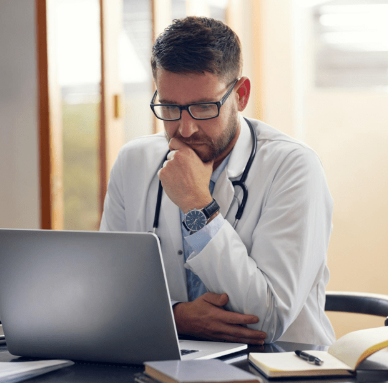 A male doctor with glasses, wearing a white coat and stethoscope, sitting at a desk and looking at a laptop, with a thoughtful expression.