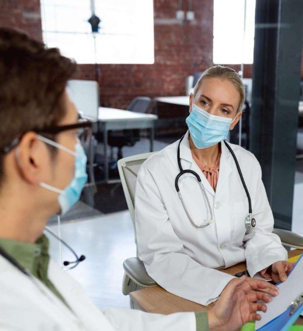 Two healthcare professionals wearing face masks, one male and one female, sitting at a table in a medical office, engaged in conversation.
