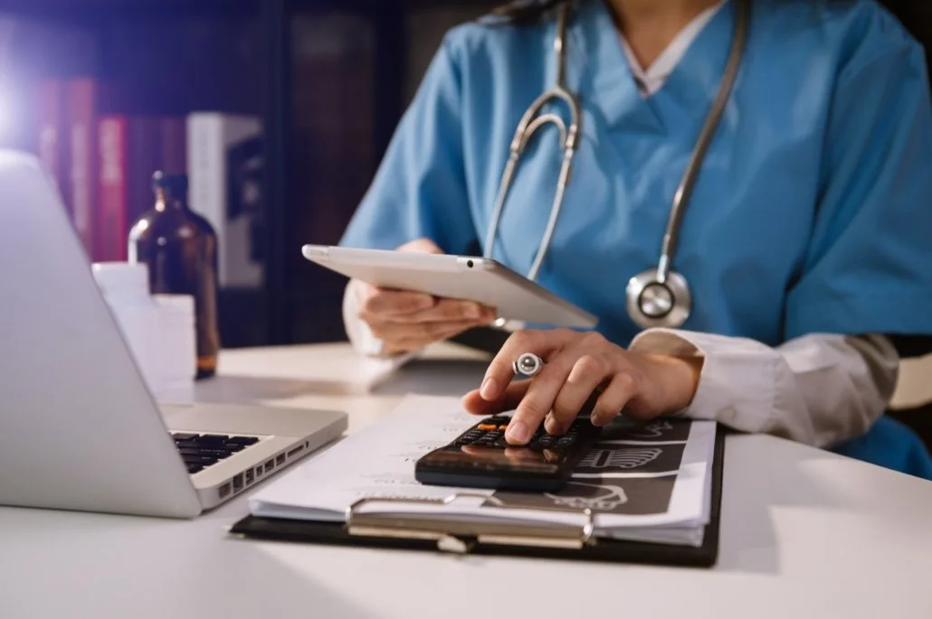 Healthcare professional in blue scrubs and stethoscope working at a desk with a laptop, calculator, tablet, and documents.