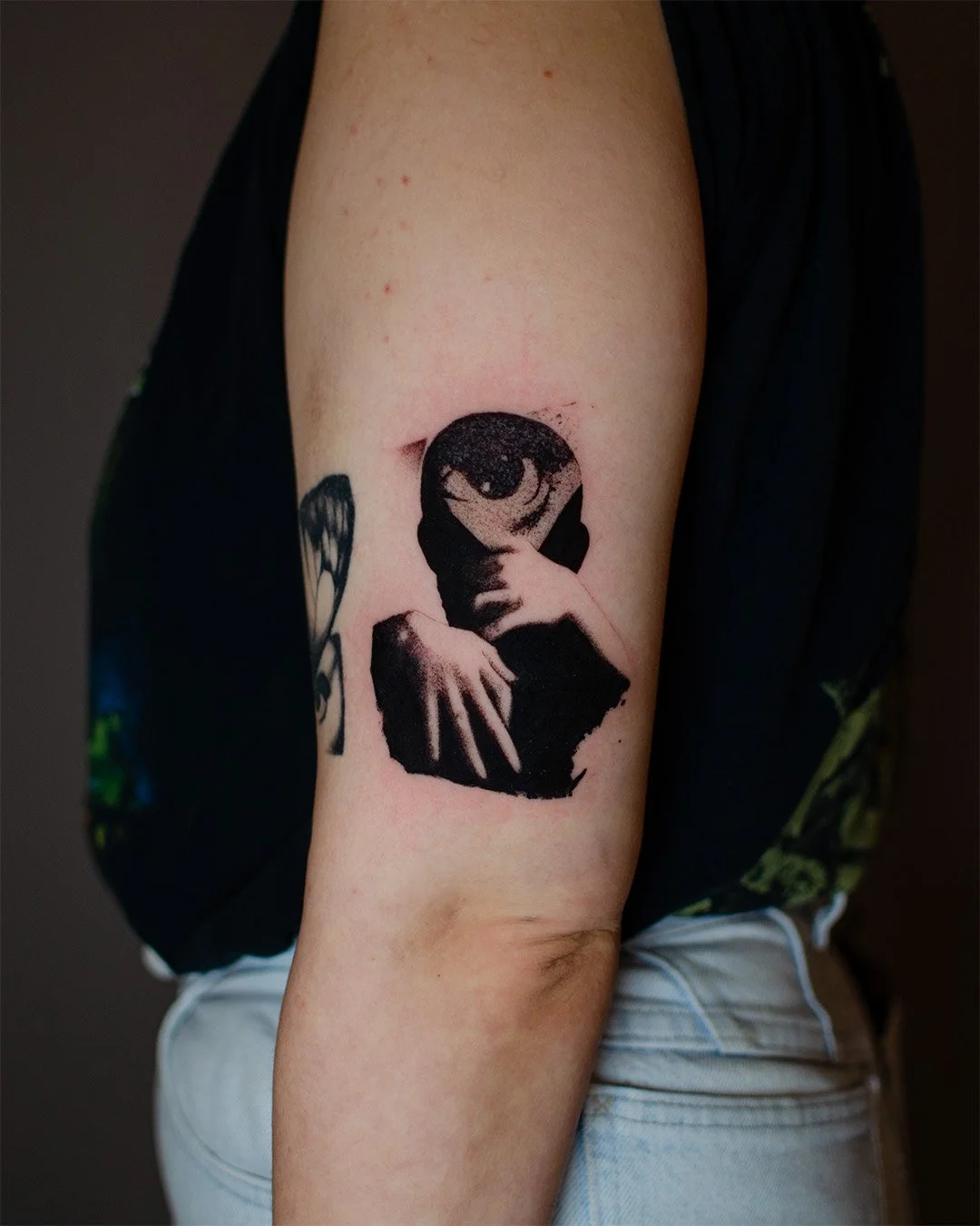 Tattoo of a person with their face partially covered by their hand, with one eye visible, on a person's upper arm.